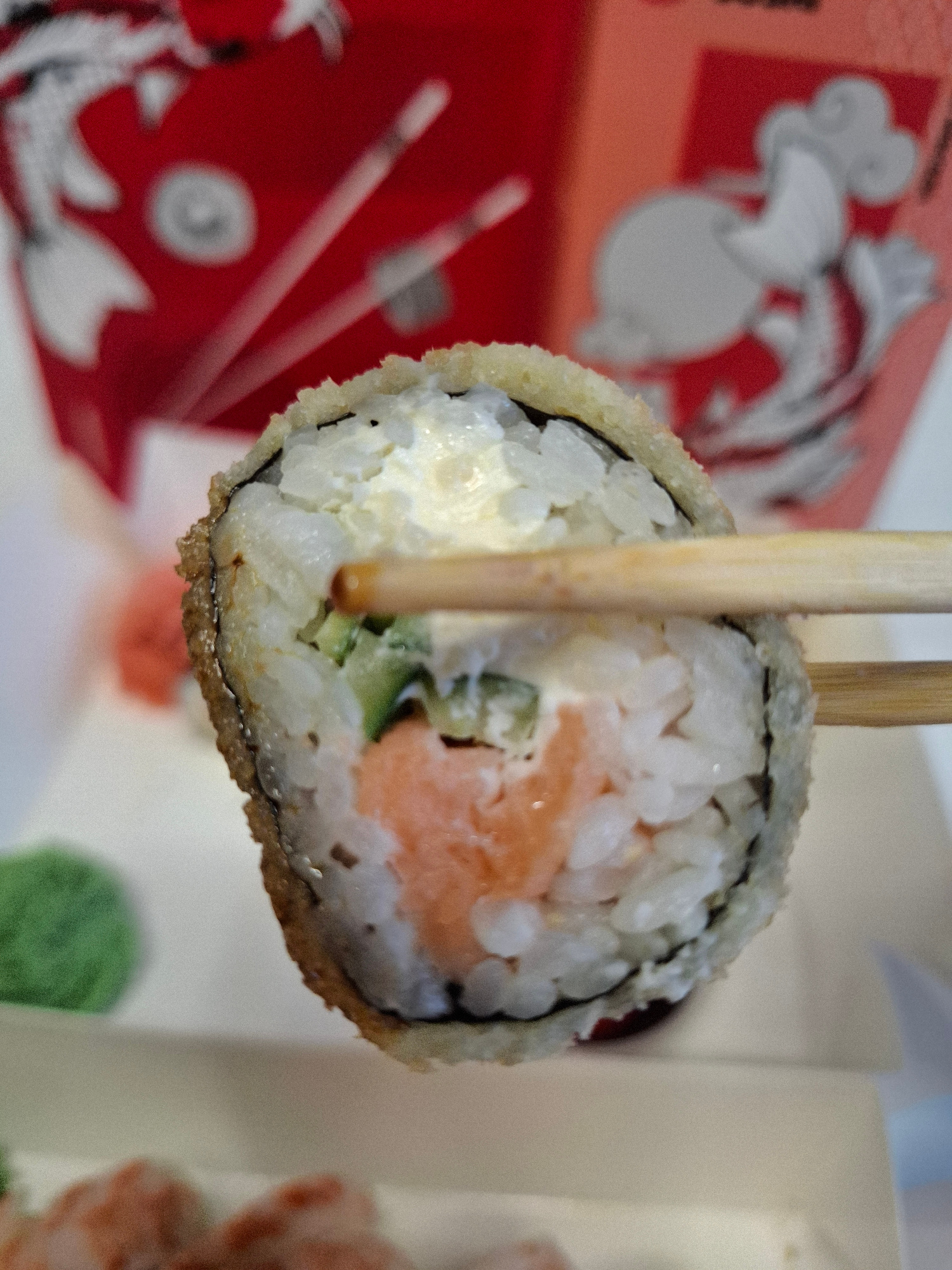 A close-up, slightly blurred shot shows a piece of sushi, likely a roll, being held by wooden chopsticks. The sushi appears to be deep-fried, with a crispy, golden-brown outer layer. Inside, layers of white rice, a pink filling (likely salmon), green cucumber, and a white creamy ingredient (possibly cream cheese or mayonnaise) are visible. In the background, a red container with white Asian-inspired motifs is partially visible. To the left and bottom of the sushi, out-of-focus green and pinkish elements suggest other food items, possibly wasabi and pickled ginger, or other sushi components, on a light-colored plate or surface. The lighting is bright, highlighting the textures of the food. The scene is devoid of any discernible landmarks or elements that would allow for the identification of a specific location within Bratislava, Slovakia.