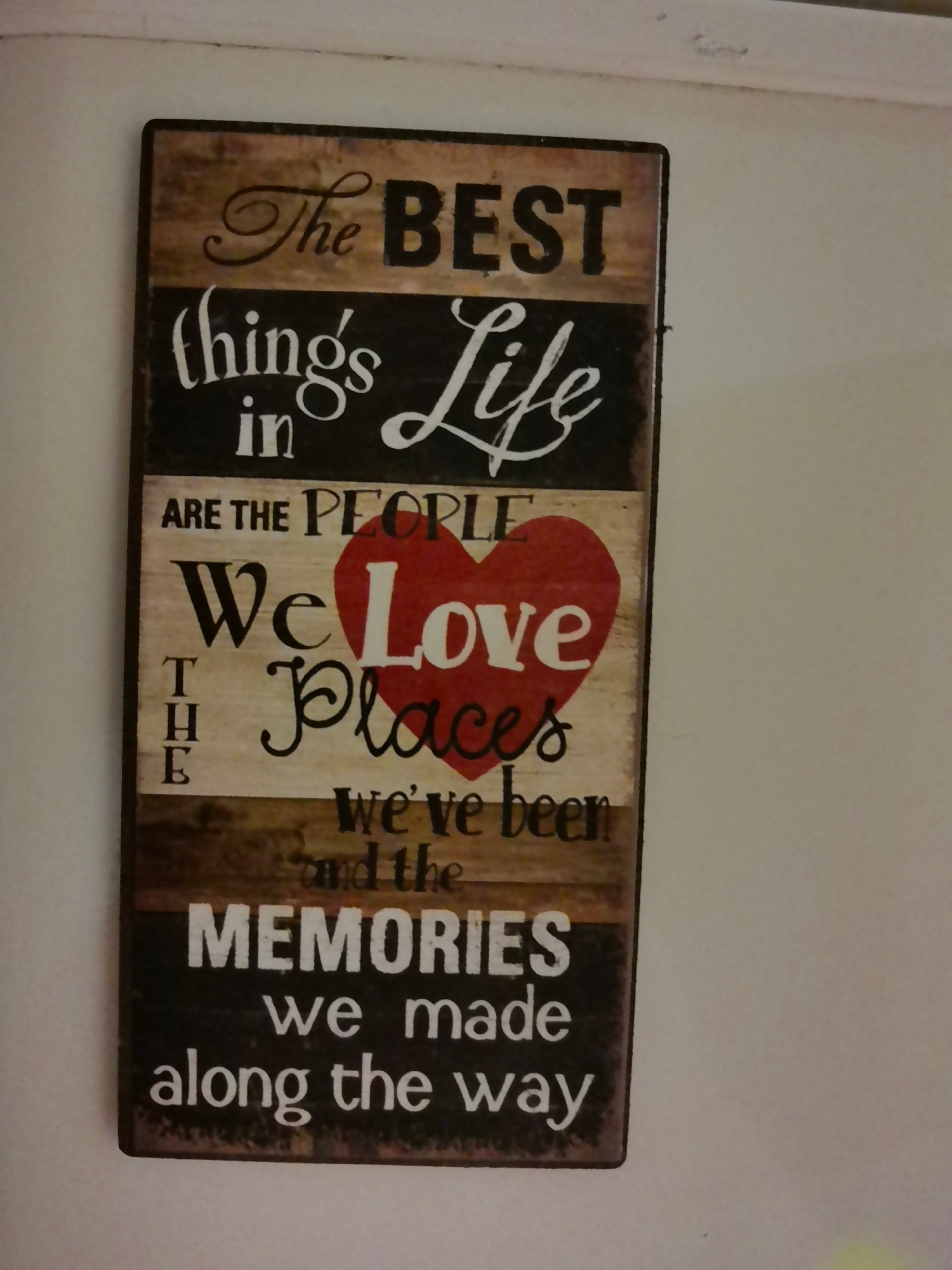 A vertical, rustic wooden sign with painted text is mounted on a plain, light-colored wall. The sign has a distressed, weathered appearance with visible wood grain and dark framing. The text on the sign reads: "The BEST things in Life ARE THE PEOPLE We Love Places we've been and the MEMORIES we made along the way." The word "Love" is prominently displayed in a large red heart shape. The sign is illuminated from the front, casting a slight shadow on the wall behind it. The environment appears to be indoors, possibly a home or a decorative display area. There are no people visible in the image. The overall impression is one of sentiment and domesticity. The location is identified as Aksakovo, Bulgaria.