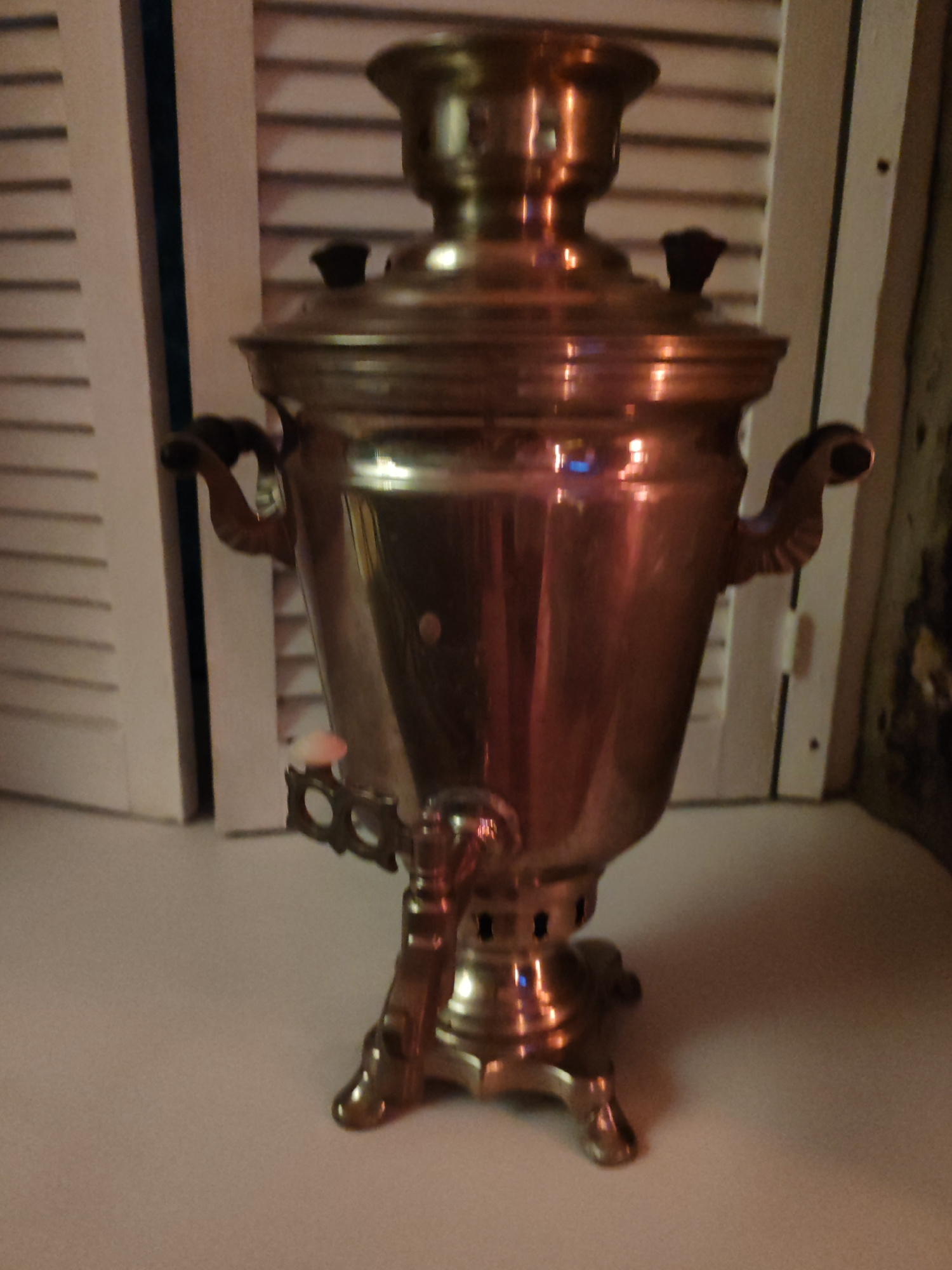 A metallic samovar, likely brass or copper, sits on a light-colored surface. The samovar has a tall, ornate body with a lid and handles. It is positioned in front of a white shuttered background. The lighting is dim, casting warm reflections on the samovar's surface. There are no people or animals present in the image. The scene appears to be indoors, possibly a domestic setting. The location in Komsomolsk-on-Amur, Russia, is not discernible from the visual information provided.