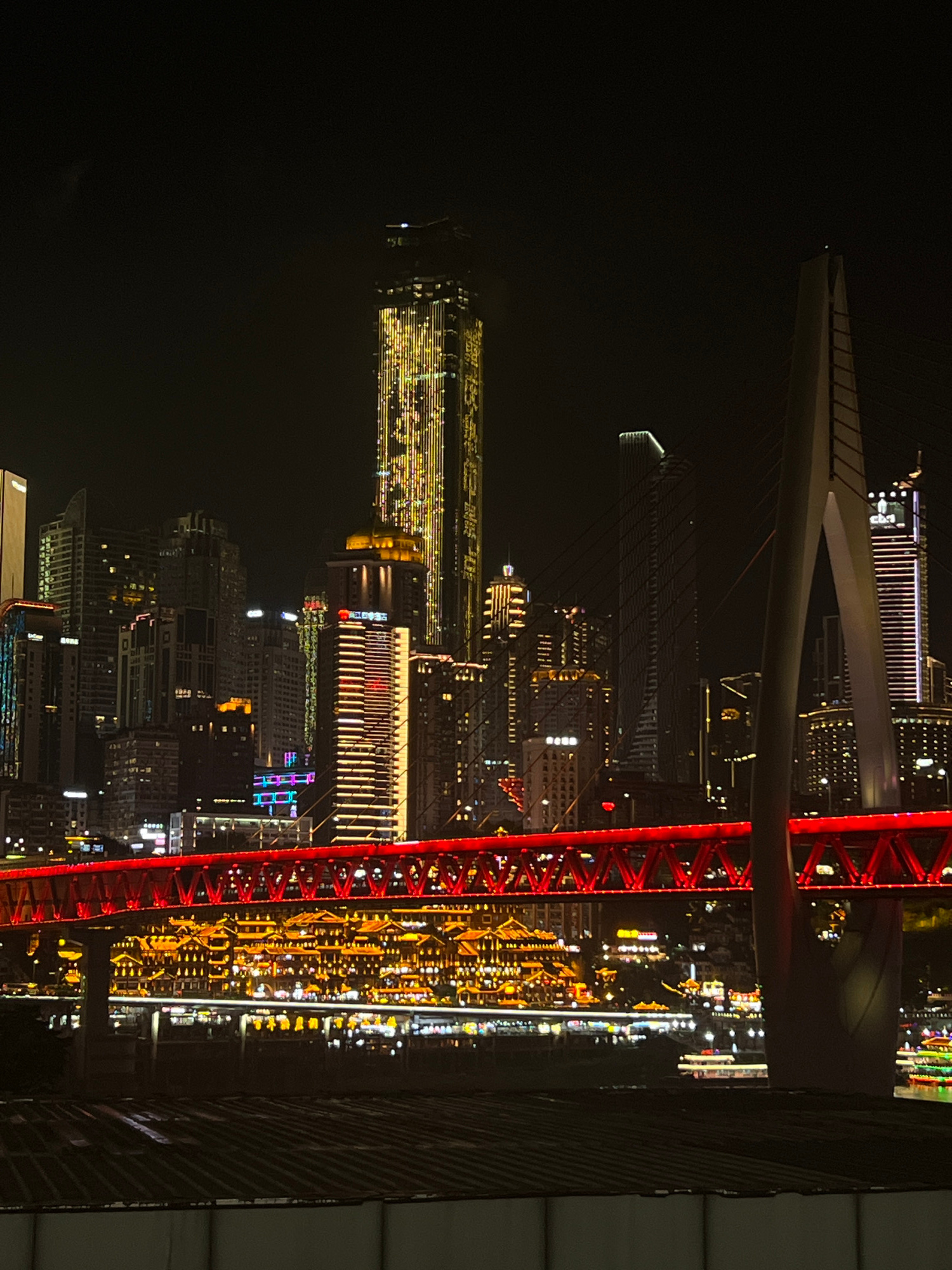 The image shows a nighttime cityscape with a prominent red bridge in the foreground. The bridge is illuminated with a vibrant red light, highlighting its intricate metal structure. Beyond the bridge, a dense collection of skyscrapers pierces the dark sky, their windows aglow with various colors of light, predominantly yellow and white. Some buildings are lit up in patterns, suggesting architectural illumination.
The scene depicts a bustling urban environment at night. The time of day is clearly nighttime, as indicated by the darkness of the sky and the artificial illumination of the city. There are no visible people or specific activities taking place, suggesting the image is a general view of the city. The setting appears to be a waterfront area, as suggested by the reflections of lights that could be on water, and the arrangement of buildings. The overall mood conveyed is one of urban vitality and modern architectural grandeur.
There is some visible text on one of the buildings in the middle distance, appearing as red characters, possibly a business name or advertisement, but it is not legible. The location context provided is Jiangbeicheng Subdistrict, China, which aligns with the appearance of a major Chinese urban center. The image is oriented vertically, with the bridge and city appearing upside down relative to a typical horizontal view.