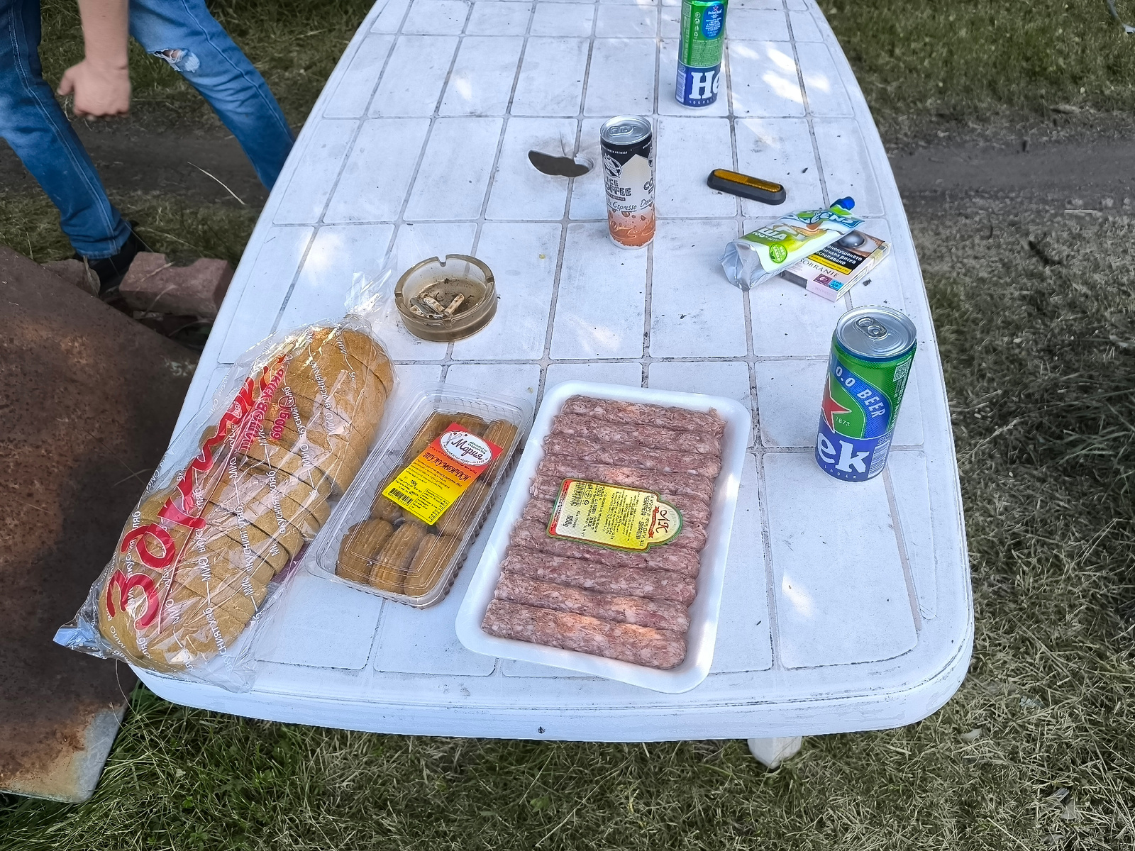 A high-angle medium shot captures a picnic scene on a white, weathered plastic table in an outdoor setting with green grass. The table is laden with food and beverages, suggesting a casual meal or gathering. Prominently displayed are a loaf of sliced bread in plastic packaging, a container of what appears to be pastry or dessert items labeled "Мария" (Maria) and "ТУЛУМБИЧКИ" (Tulumbichki), and a tray of raw meat, likely sausages or kebabs, with Cyrillic text on a yellow label.
Several drinks are also visible: a can of "0.0 BEER" and another can with "H" and "Espresso Do" printed on it, suggesting coffee. A pack of cigarettes labeled "SOBRANIE" and an ashtray filled with cigarette butts indicate smoking. A person's legs, clad in ripped blue jeans, are visible in the upper left corner, with one hand reaching towards something out of frame, hinting at an ongoing activity. The presence of the food items and drinks, along with the person's attire and relaxed setting, points towards a social gathering like a barbecue or picnic. The lighting suggests it is daytime, possibly late afternoon or early evening, with natural light illuminating the scene. The overall mood appears to be relaxed and informal.