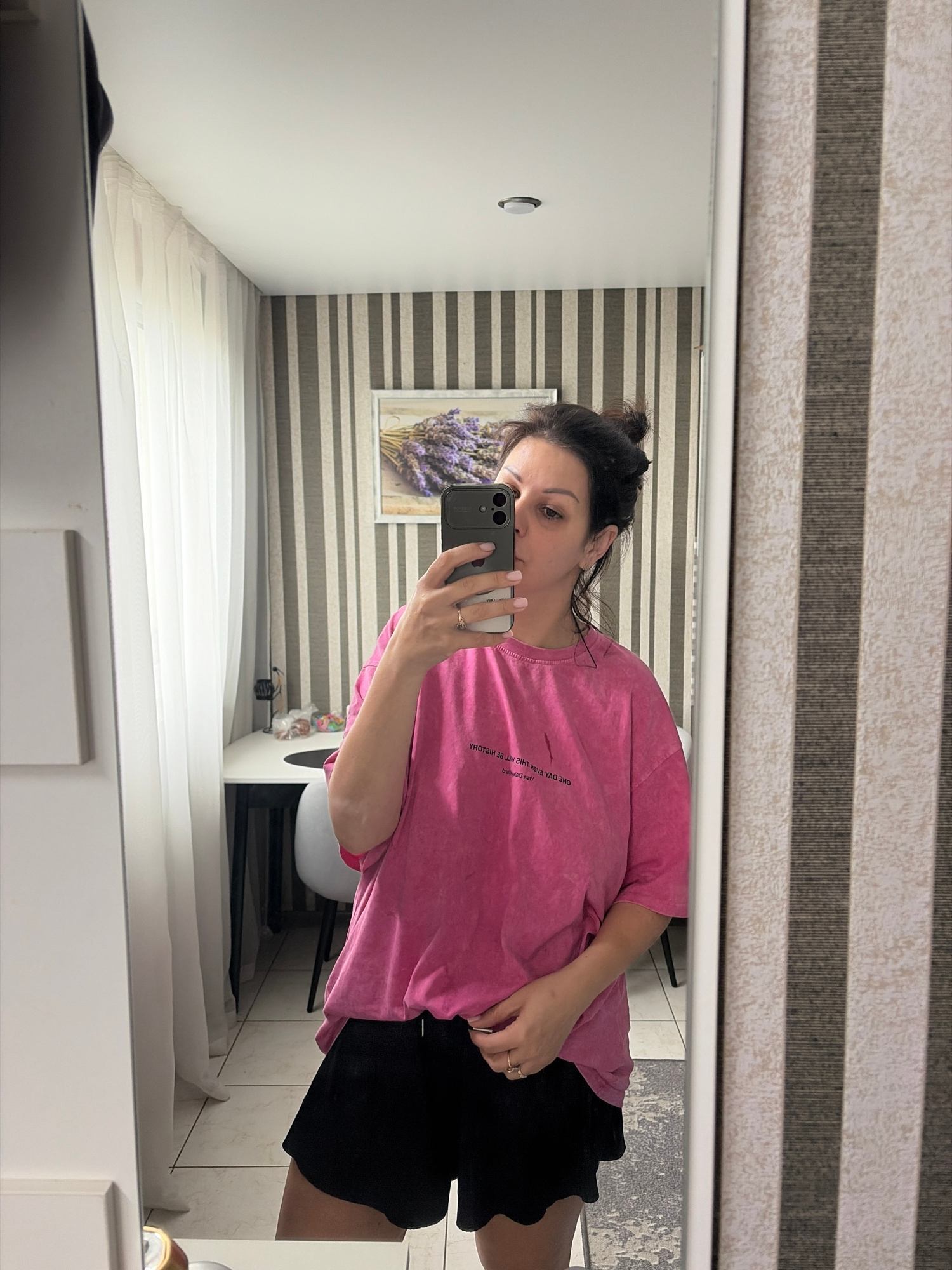 A woman with her dark hair pulled up in a bun takes a mirror selfie in a well-lit room. She is wearing a pink oversized t-shirt with text and black shorts. She is holding a smartphone in her right hand to take the picture. The background includes a wall with vertical striped wallpaper, sheer white curtains, and a framed picture of lavender. The floor is tiled. A table and chairs are visible in the reflection of the mirror. The overall impression is one of a casual indoor setting, possibly a dressing room or bedroom.