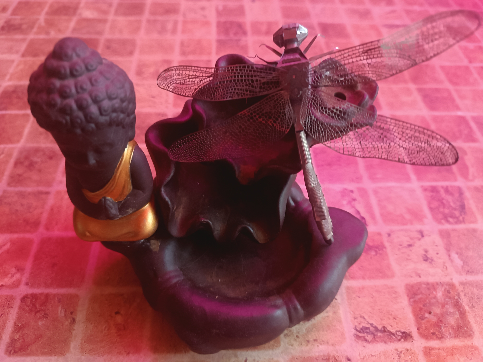 The image shows a small, dark-colored Buddha figurine sitting on a dark, sculpted base that resembles a lotus flower. A metallic dragonfly ornament is perched on the base in front of the Buddha. The figurine and ornament are placed on a pink, tiled surface. There are no people present in the image. The image does not contain any information about the city of Sluck, Belarus, or any other location.
