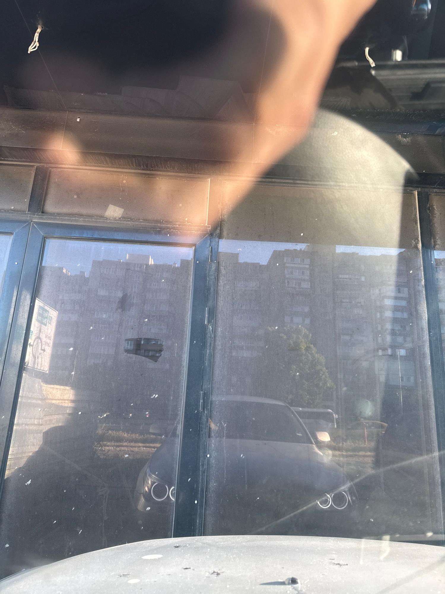 The image shows a reflection on a dusty glass surface, likely a storefront or window in Varna, Bulgaria. The reflection depicts an outdoor scene with several apartment buildings in the background, appearing somewhat weathered. In the foreground of the reflection, a dark-colored car with distinctive circular headlights is visible. Sunlight casts a strong glare across the glass, obscuring some details and creating bright highlights. A human hand is also partially visible in the upper right corner of the image, casting a shadow on the glass, suggesting the photograph was taken from inside a vehicle or from a position close to the glass. The overall impression is a slightly hazy, urban environment.