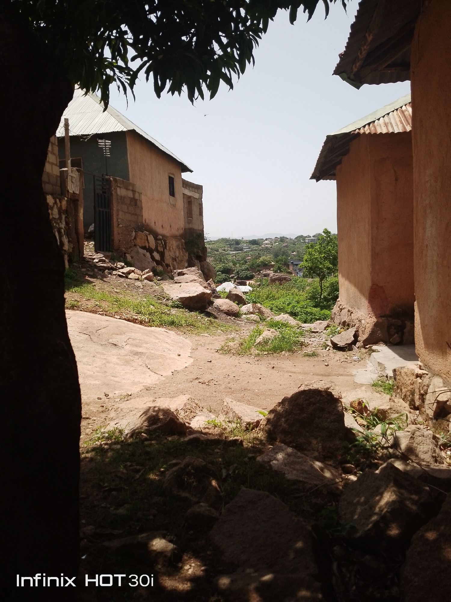 A medium shot captures a view down a rocky, dirt path between two buildings. The building on the left is made of stone and has a green, stuccoed facade with a corrugated metal roof. A metal gate is visible in an opening in the wall. The building on the right is made of reddish-brown stucco with a corrugated metal roof. In the distance, a lush green valley is visible with scattered houses and trees under a hazy sky. The foreground is filled with rocks and sparse vegetation, and a large tree trunk is partially visible on the left. The bottom of the image contains the text "Infinix HOT 30i".