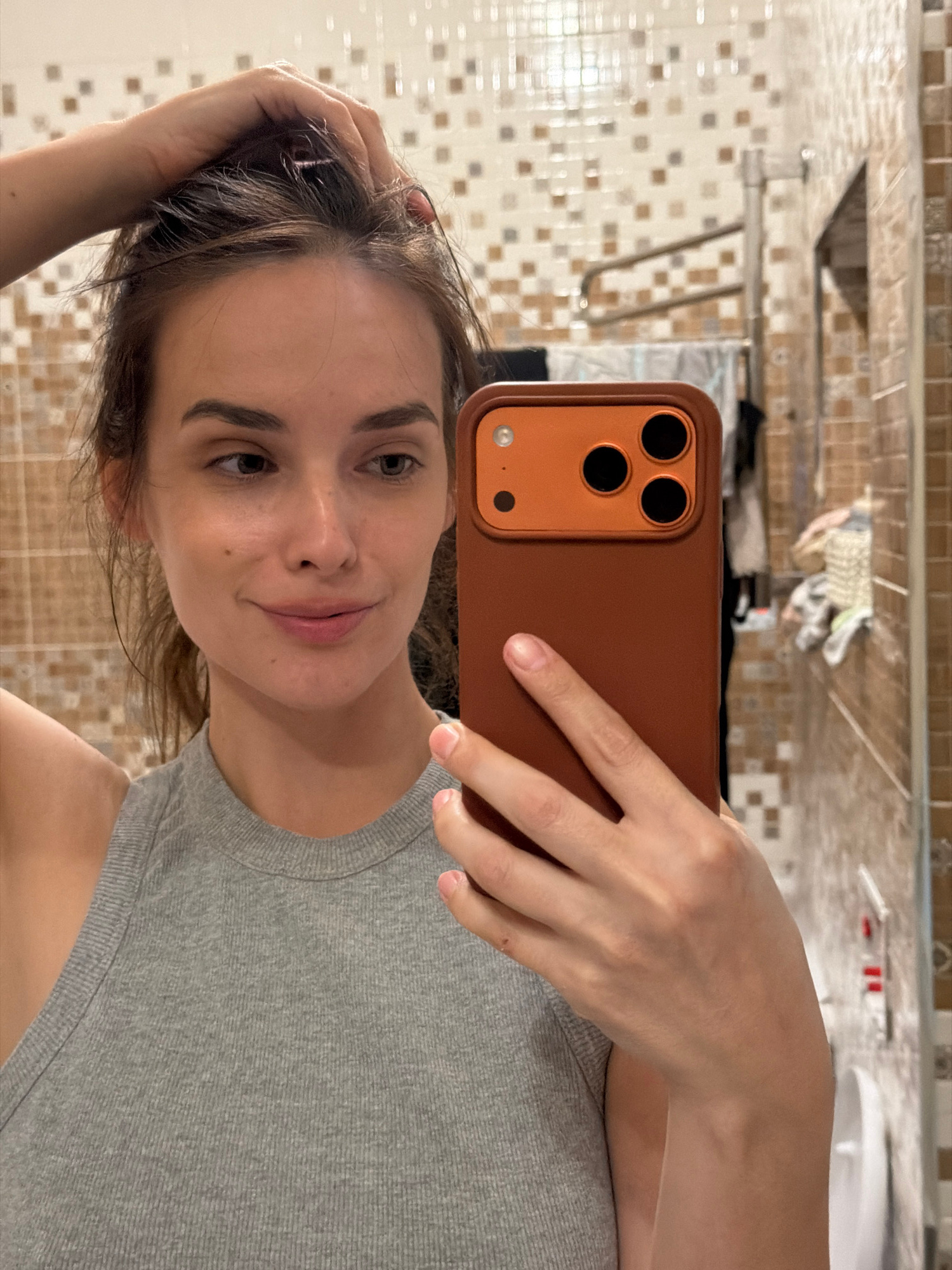 A young woman with brown hair and brown eyes is taking a selfie in a bathroom. She is wearing a grey tank top and is holding an orange phone in her right hand. Her left hand is in her hair. The bathroom has tiled walls with a checkered pattern of brown, beige, and white squares. A towel rack is visible in the background, with towels hanging on it. The overall lighting is dim, typical of an indoor environment.