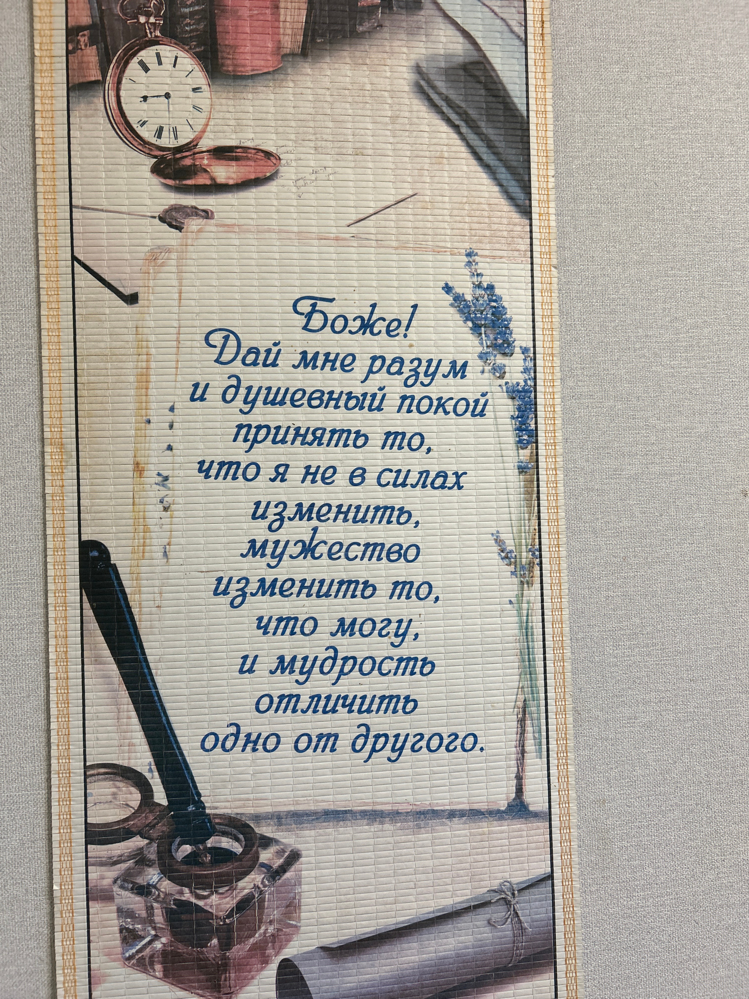 The image depicts a woven mat with a prayer written in Cyrillic script. The prayer reads: "Боже! Дай мне разум и душевный покой принять то, что я не в силах изменить, мужество изменить то, что могу, и мудрость отличить одно от другого." This translates to: "God! Grant me the wisdom and peace of mind to accept what I cannot change, the courage to change what I can, and the wisdom to know the difference."
The mat features decorative elements including an antique pocket watch, an inkwell with a quill pen, a sprig of lavender, and rolled documents. The overall aesthetic is vintage and reminiscent of a scholar's desk. The mat is placed on a textured, light-colored surface, likely a wall or furniture.
There is no information in the image to suggest a specific location in the city of Yakutsk, Russia. The scene is entirely internal and decorative. There are no people present in the image.