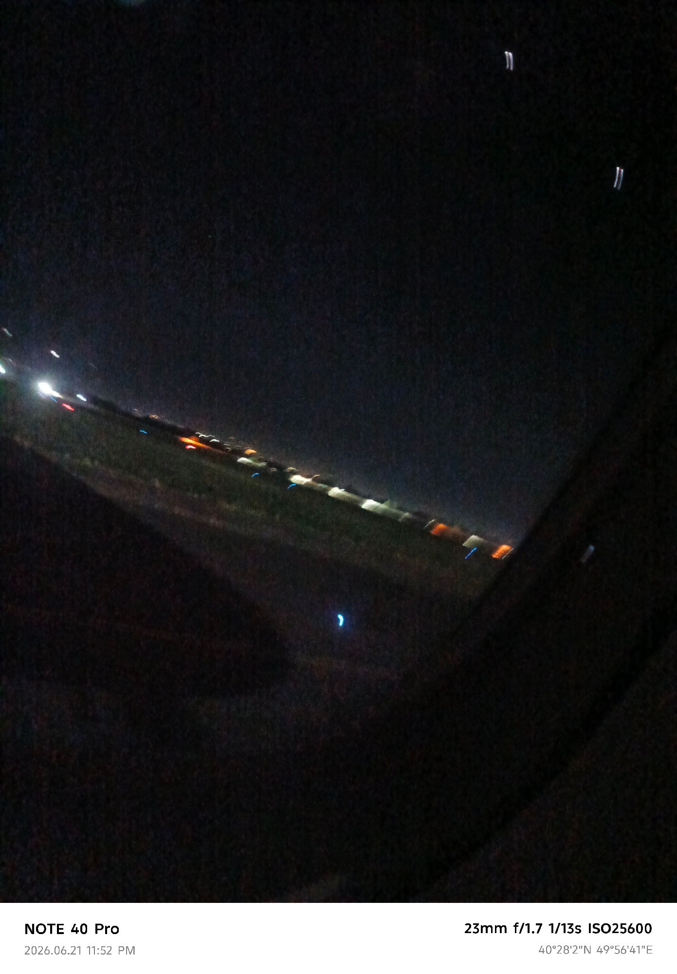 The image captures a night view from an elevated position, likely an airplane, overlooking a city at dusk or night. Streaks of light indicate moving vehicles on a road, suggesting an urban environment. The lights are blurred due to the long exposure (1/13s) and the movement of the camera. The scene is dark with scattered artificial lights illuminating the landscape. The text overlay indicates the photograph was taken with a "NOTE 40 Pro" at "2026.06.21 11:52 PM" with settings "23mm f/1.7 1/13s ISO25600". The GPS coordinates provided are "40°28'2''N 49°56'41''E". These coordinates correspond to a location in Azerbaijan, not Dushanbe, Tajikistan. Therefore, the description will focus on the visual elements of the image without referencing Dushanbe.
