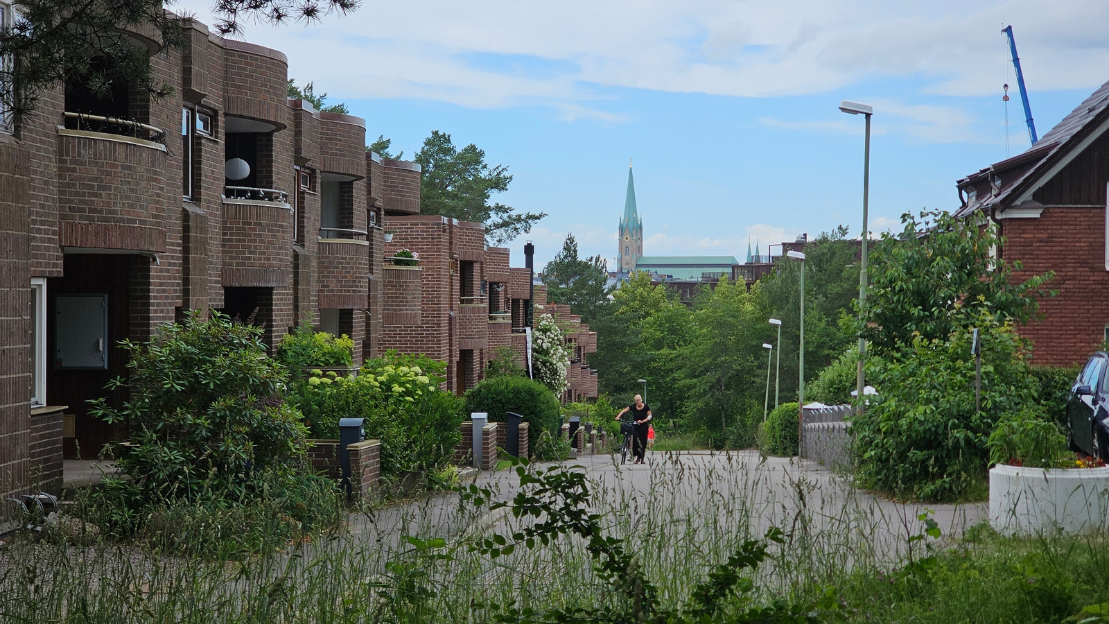 The image depicts a street scene in Linköping, Sweden. In the foreground, tall grass lines a paved road. On the left side of the road, a multi-story building with a distinctive brick facade and curved balconies is visible. This building appears to be residential, with several windows and balconies adorned with plants. On the right side of the road, a residential house with a red brick exterior is partially visible, along with a dark-colored car parked in front of it.
Further down the road, two individuals are seen walking. One person is on a bicycle, wearing dark clothing and pushing it along the road, while another person in black clothing walks alongside. In the background, a dense canopy of green trees forms a lush backdrop. Beyond the trees, the spire of a prominent church, likely a landmark in Linköping, rises into the sky, accompanied by other buildings and structures. A blue construction crane is also visible in the distance, indicating ongoing development in the city. The sky is partly cloudy, with a mix of blue sky and white clouds. The overall impression is a suburban or semi-urban environment with a blend of residential architecture, natural greenery, and distant city landmarks.