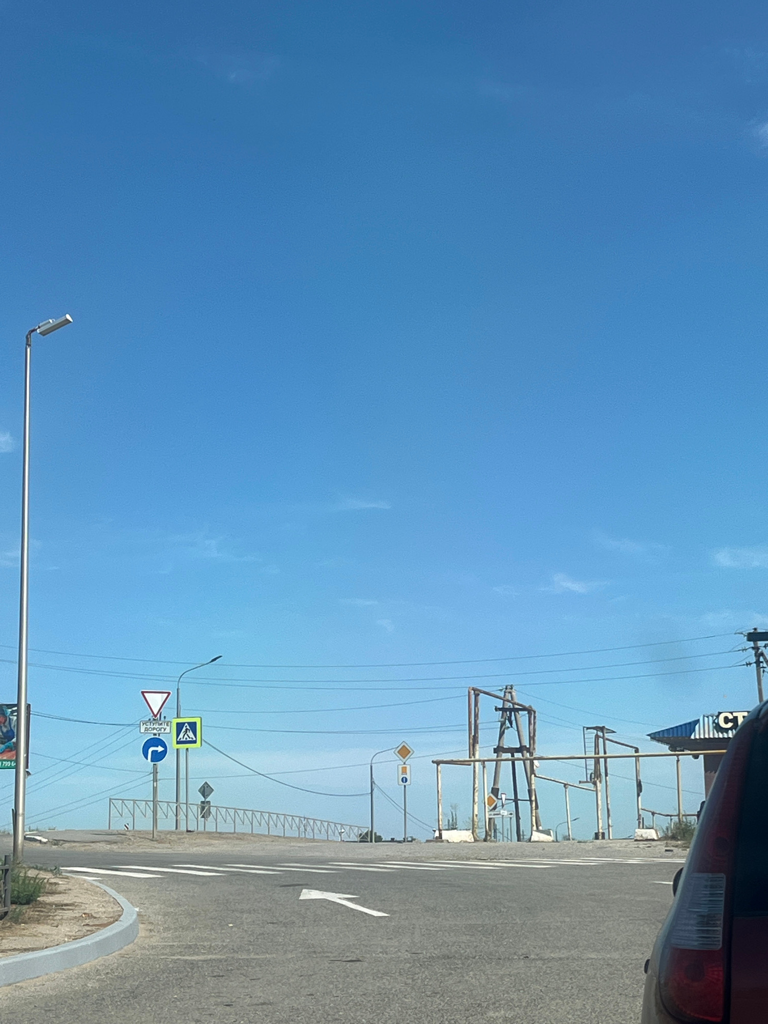 The image shows a view from a vehicle, looking towards a road intersection under a clear blue sky. The road surface is asphalt, with white lane markings and a pedestrian crossing. There are several road signs visible, including a yield sign, a pedestrian crossing sign, and a sign indicating a right turn. A street lamp is present on the left side of the image. In the background, there are structures that appear to be part of an industrial or utilitarian facility, with metal frames and supports. A building with a blue roof and the Cyrillic letters "СТ" is also visible. The presence of these elements and the overall aesthetic suggest a location in a rural or semi-urban area in Russia, potentially Ikryanoye, Russia, given the context. The overall scene depicts a typical road junction in daylight.