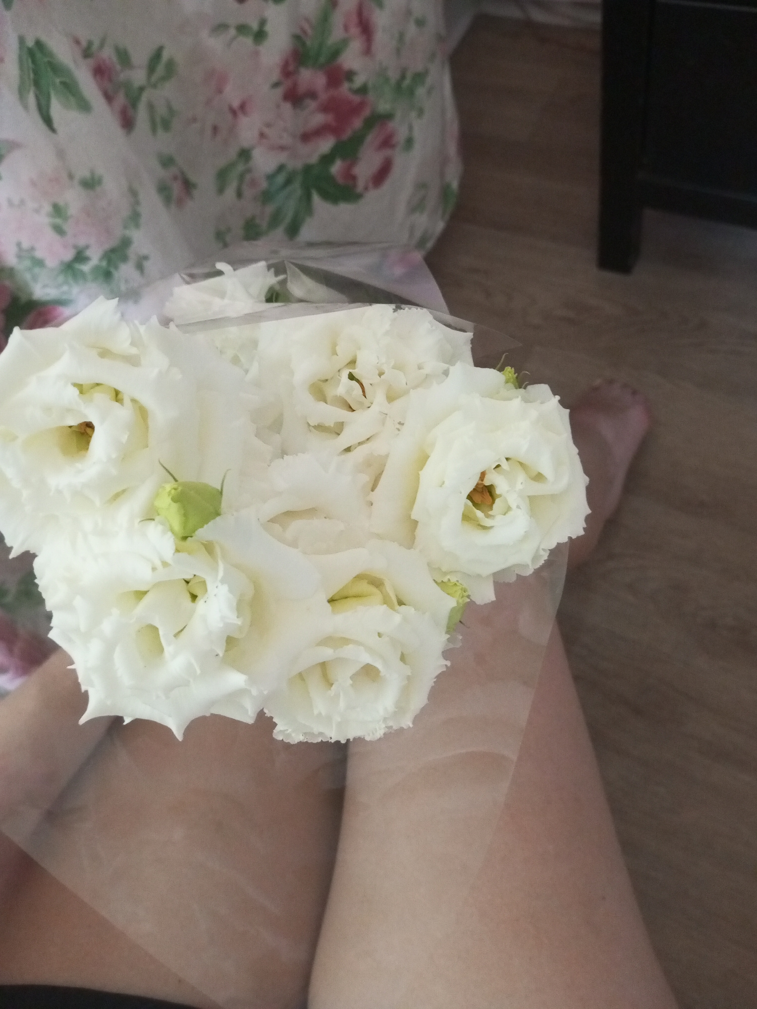 A bouquet of white roses is held by a person whose legs are visible. The roses are in full bloom with delicate, ruffled petals and some have a faint yellow or orange center. There is a small, light green bud amongst the flowers. The background consists of a wooden floor and a piece of fabric with a floral pattern featuring pink and green flowers. A portion of a dark piece of furniture is also visible on the right side of the image. A person's bare foot is visible on the wooden floor behind the bouquet. The scene appears to be indoors.