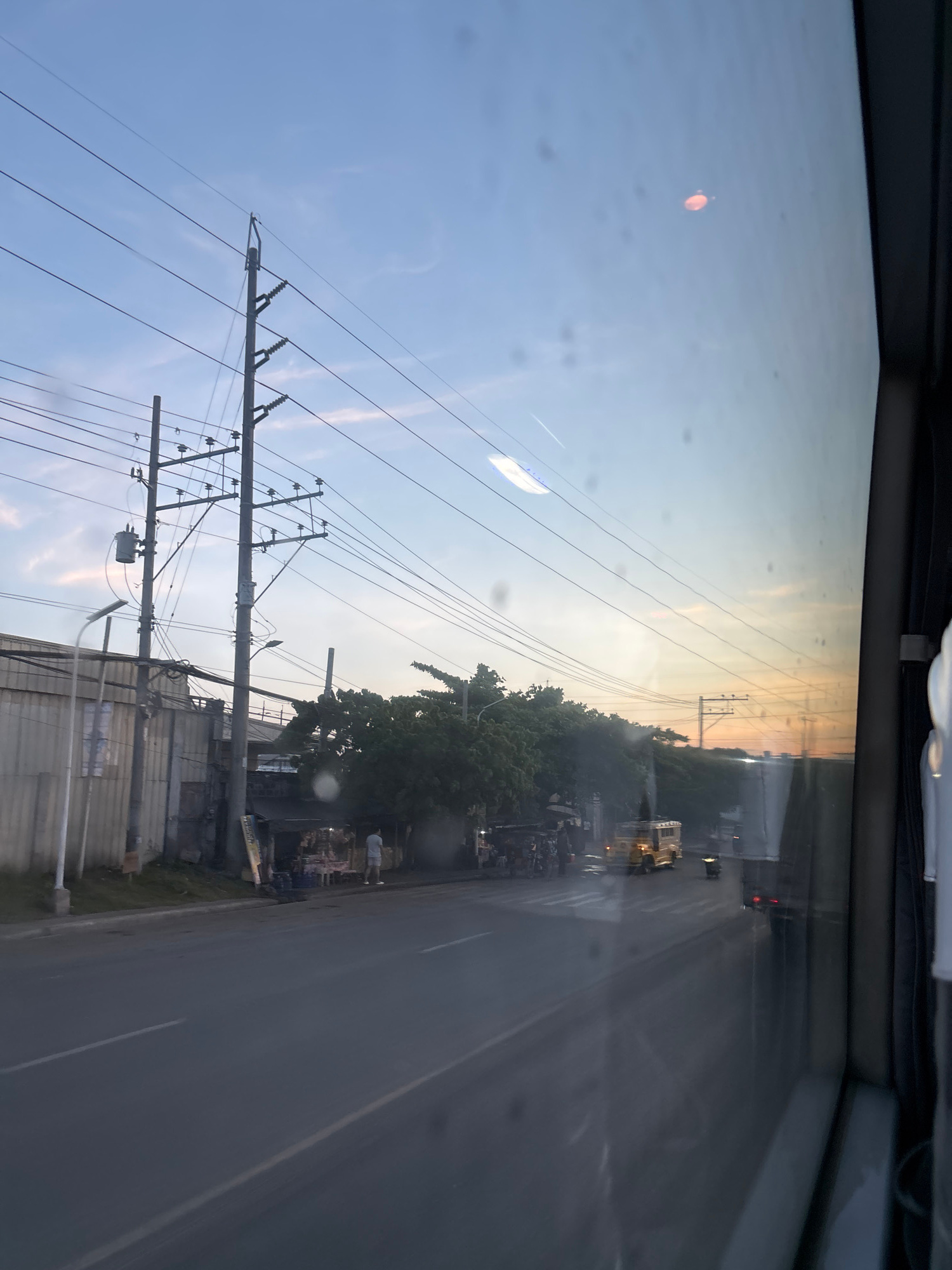 The image shows a view from inside a vehicle, looking out through a dirty window at a road and its surroundings at dusk in Davao City, Philippines. On the left, there is a light-colored wall with some signs and a streetlamp. A series of utility poles with wires and transformers are prominent in the scene, stretching across the sky. Along the roadside, there are some small stalls or shops with awnings, and a few people are visible standing near them. A yellow jeepney is driving on the road, and another vehicle is behind it. Trees line the right side of the road, and the sky above is a gradient of blue, transitioning to an orange and yellow hue near the horizon, indicating sunset. The window has several water spots or smudges.