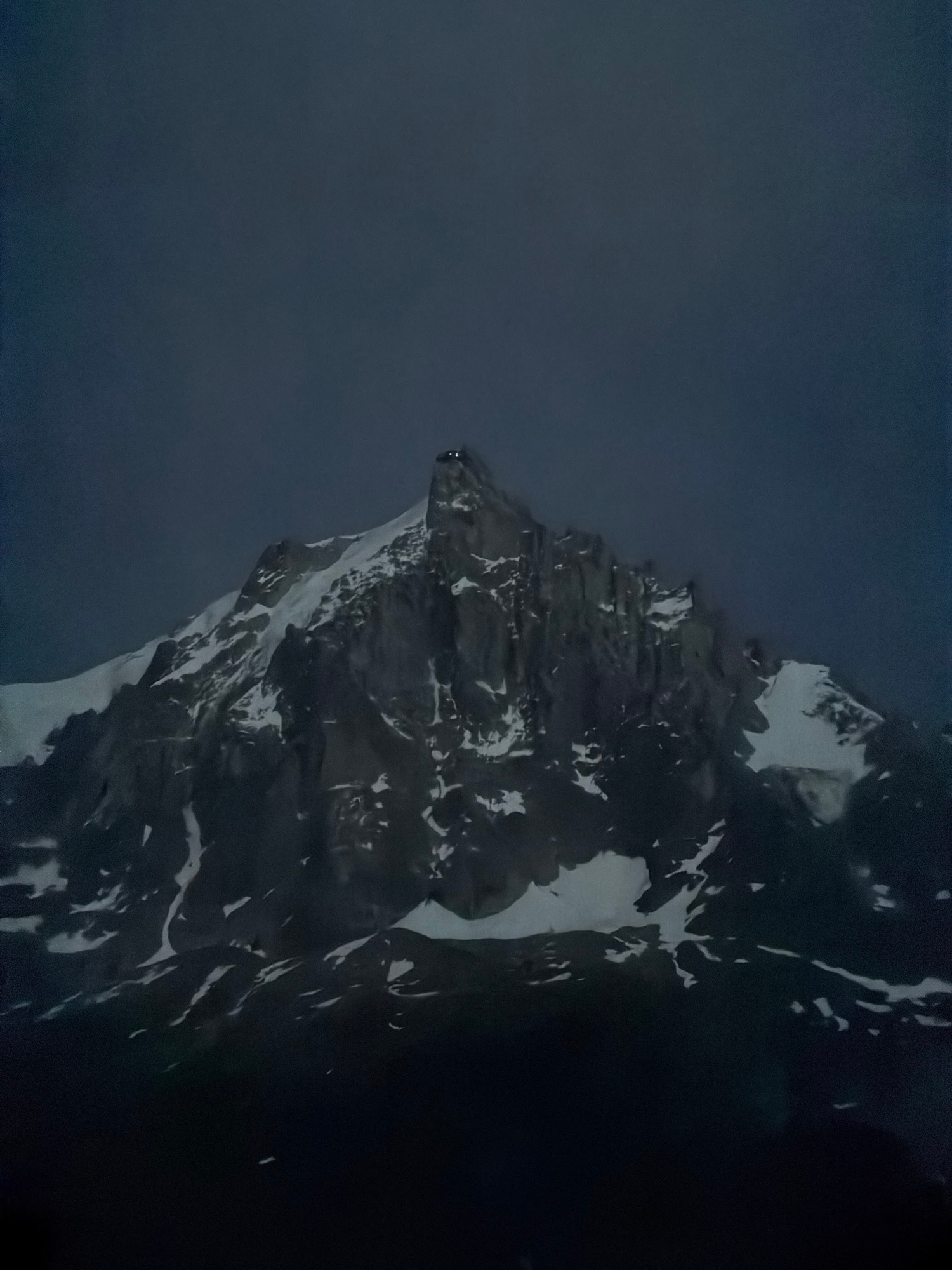 The image depicts a dramatic, snow-capped mountain peak silhouetted against a dark, overcast sky. The mountain's rugged, rocky face is partially covered in snow, creating stark contrasts of light and shadow. The lower portion of the image fades into darkness, obscuring the base of the mountain and any surrounding landscape. The overall impression is one of imposing natural grandeur and a somewhat somber, moody atmosphere. This could be a view of Mont Blanc from Chamonix-Mont-Blanc, France, given the iconic mountain range in the area.
