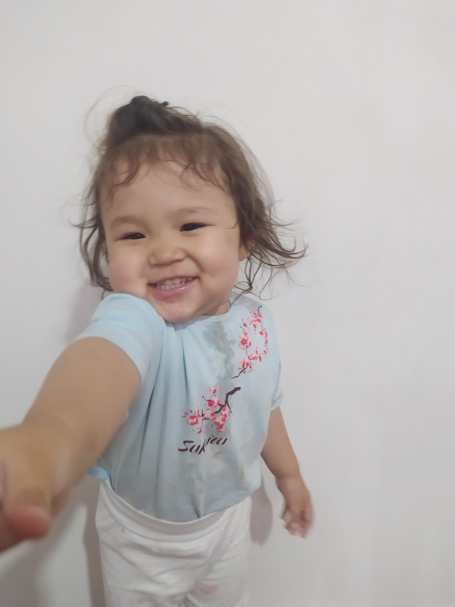 This is a portrait-oriented, horizontally oriented, medium shot of a young child lying on a white surface, possibly a bed or floor. The child, appearing to be a toddler, has dark, wavy hair and a fair complexion. They are looking towards the camera with a wide, happy smile, showing visible teeth. The child's eyes are dark and appear to be looking slightly to the left of the camera.
The child is wearing a light blue t-shirt with a pink cherry blossom design and the word "Sakura" written in dark lettering. Below the t-shirt, white fabric is visible, suggesting they are also wearing white shorts or pants. The child's right arm is extended towards the camera, with the hand blurred due to motion.
The background is a plain, light-colored wall, creating a simple and uncluttered setting. The lighting appears to be soft, possibly natural light from a window, casting subtle shadows on the white surface. The overall mood is joyful and innocent, conveyed by the child's bright smile. There are no clear indicators of the time of day, weather, or specific location beyond the provided context.