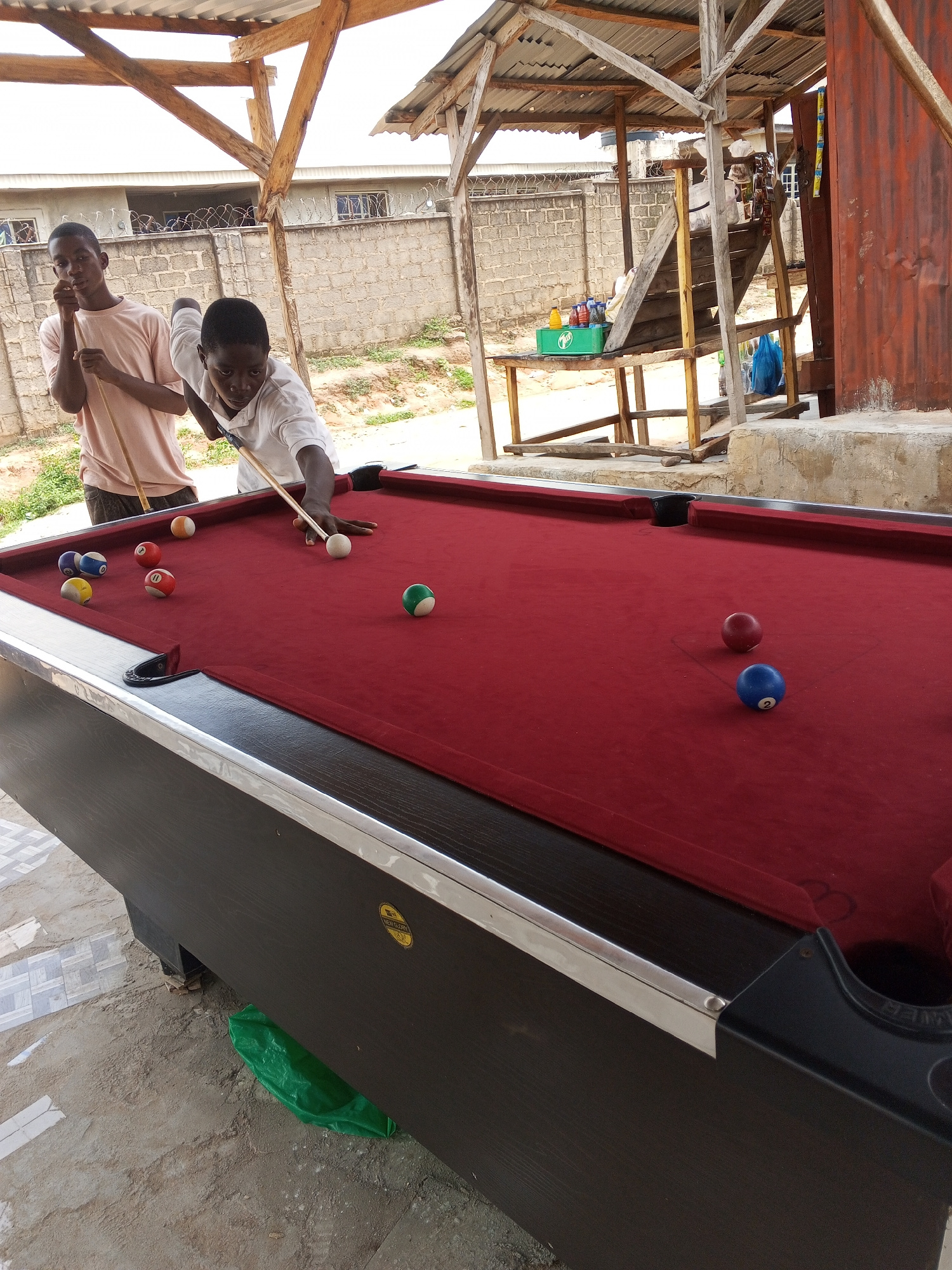 Two young men are playing billiards on a red felt pool table under a makeshift wooden shelter. The shelter has a corrugated metal roof and open sides, revealing a brick wall and some vegetation in the background. The ground around the pool table is uneven and appears to be dirt and concrete. The man on the right is in the process of striking the cue ball with his cue stick, aiming for another ball on the table. Several other billiard balls of various colors and numbers are scattered on the table. The scene is likely outdoors, possibly in a community space or an informal recreational area in Lokoja, Nigeria.
