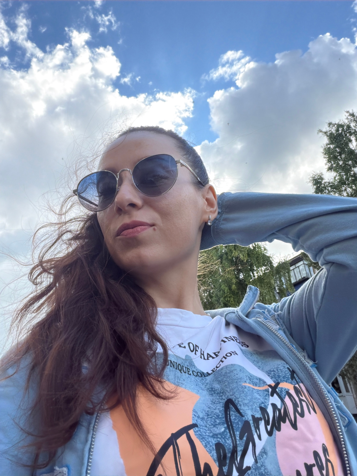 A young woman, facing upwards, is the subject of this outdoor, close-up selfie. She is wearing sunglasses and a light blue zip-up hoodie over a white t-shirt with a colorful graphic print featuring abstract shapes and black scribbled lines. The t-shirt has text that appears to say "COLLECTION OF UNIQUE" and "HANDMADES".
The background is a bright sky filled with fluffy white clouds, suggesting fair weather. Sunlight is visible, casting a glare in the upper right portion of the image. The perspective is from below, looking up at the woman. The scene appears to be outdoors, possibly in a park or residential area, with hints of trees and a building visible in the distance. The overall mood is casual and relaxed, likely captured during a daytime outing.
