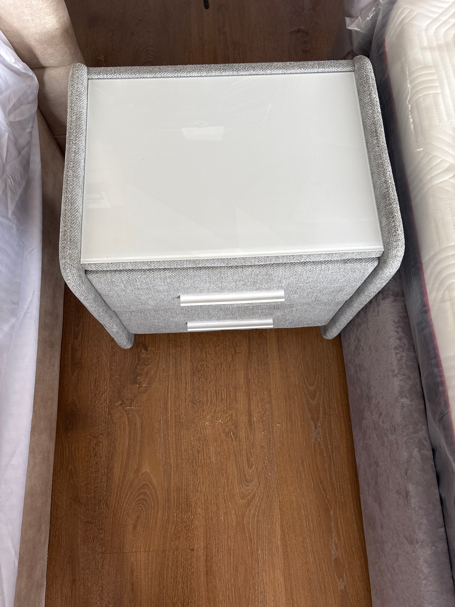 The image shows a close-up, overhead view of a bedside table next to a bed, suggesting a bedroom setting. The bedside table has a light grey, textured fabric exterior and a white, glossy drawer front with two silver handles. Above the table, a white mattress, possibly with a patterned cover, is visible, partially wrapped in plastic. A grey fabric upholstered bed frame is also visible. The surface next to the bed is a wooden floor with a warm, brown tone and distinct grain patterns. The lighting appears to be artificial, likely from an indoor source, and there are no visible indications of time of day or weather. The scene is static, with no activity taking place. The location context of Zaporizhzhia, Ukraine, does not offer any specific visual cues in this image. There is no visible text in the image.