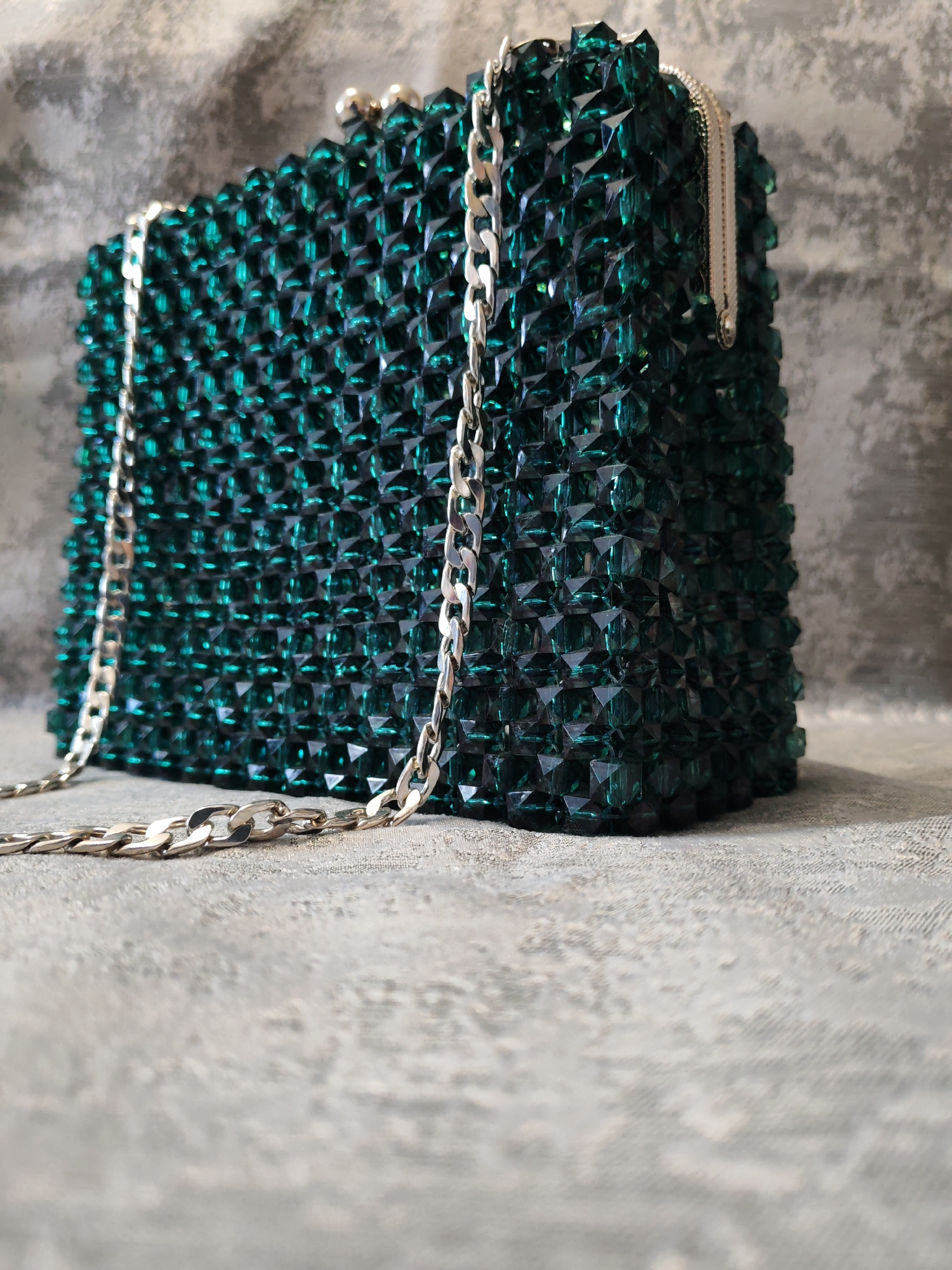This is a close-up, eye-level, medium shot of a rectangular, emerald green beaded clutch purse with a silver chain strap. The purse is adorned with numerous small, faceted green beads that create a textured, geometric surface. A silver curb chain strap is attached to the left side of the purse and drapes across the front. The purse is positioned on a textured, light grey surface that appears to be fabric or concrete, with a darker, mottled grey background. The lighting is soft, highlighting the facets of the beads and creating subtle shadows. The image is focused on the purse and its immediate surroundings, with the background slightly out of focus. There are no people present in the image. The location in the city of Сокол, Russia is not determinable from the visual information provided.