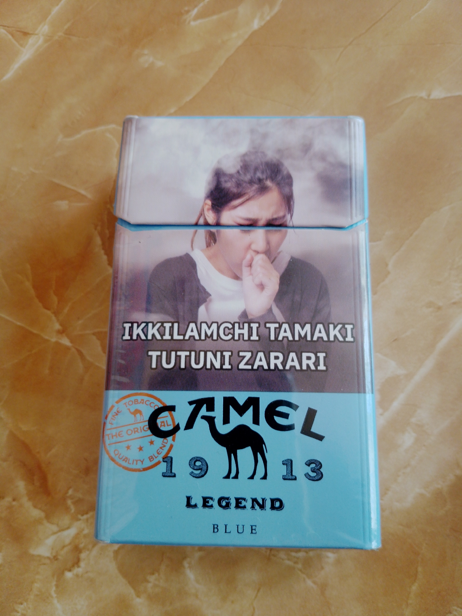 A cigarette pack is placed on a marble-patterned surface. The pack features an image of a young woman coughing, with smoke obscuring the background. Text on the pack reads "İKKİLAMÇİ TAMAKİ TUTUNİ ZARARİ" in large white letters, followed by "CAMEL" in a stylized font, a camel silhouette, "19" and "13". Below this, "LEGEND" and "BLUE" are printed. A circular emblem on the left side of the pack contains the text "FINE TOBACCO", "THE ORIG", "QUALITY", and "BLEND", with a camel graphic. The lighting is even, and the focus is on the cigarette pack. No specific city or location is identifiable from the image itself.