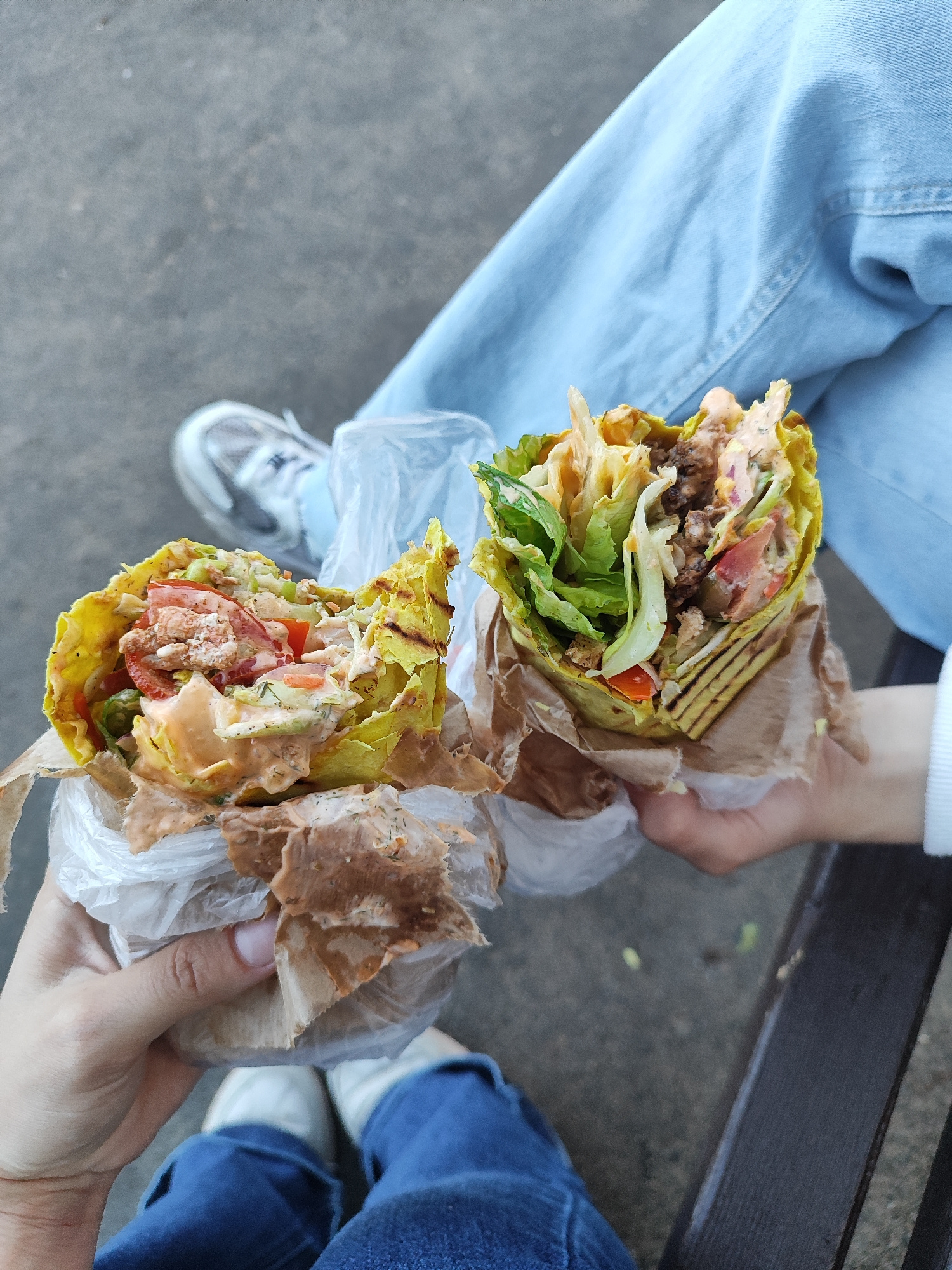 Two hands hold up two fully-stuffed shawarma wraps. The shawarma on the left is wrapped in a yellow tortilla and filled with lettuce, tomatoes, cucumbers, and a creamy sauce. The shawarma on the right is also wrapped in a yellow tortilla and appears to contain similar ingredients, with visible chunks of meat and vegetables. Both wraps are partially covered in plastic and brown paper. In the background, a person wearing blue jeans and white sneakers is visible, with legs and feet suggesting they are sitting on a dark surface, possibly a park bench. The ground beneath them is grey and textured, resembling pavement or a worn path. The scene appears to be outdoors, potentially in a public area within the city of Svietlahorsk, Belarus.