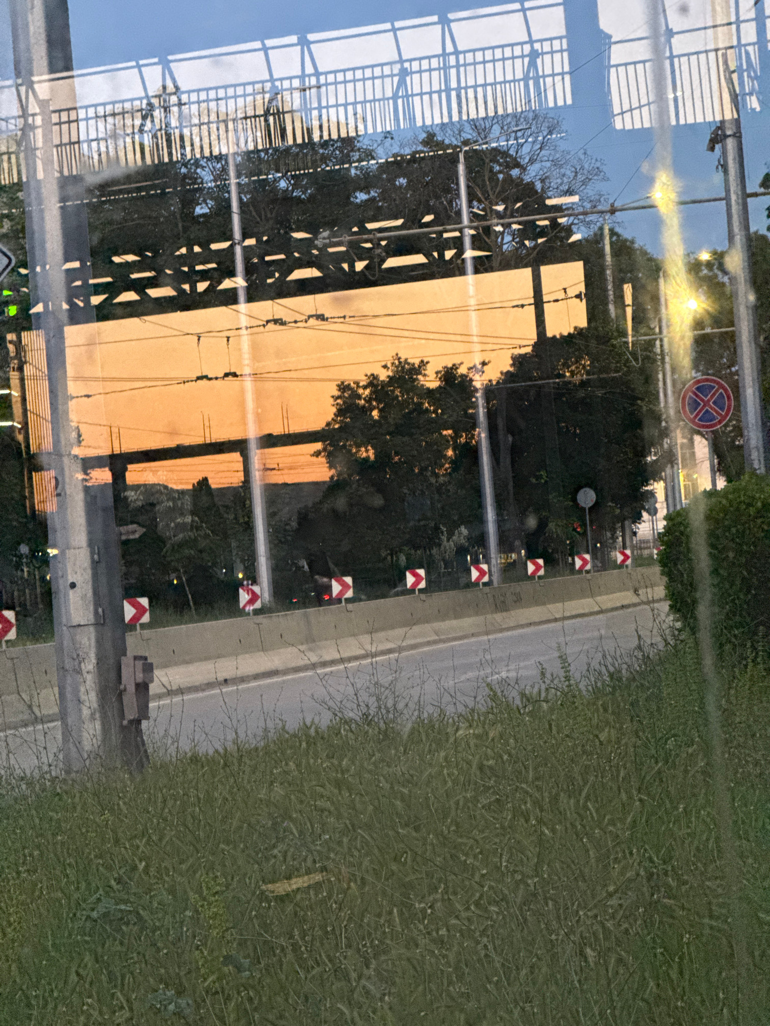 The image captures a street scene through a reflective surface, possibly a window. In the foreground, tall green grass grows along the side of a road. A concrete barrier runs alongside the road, adorned with several red and white chevron signs indicating a curve or hazard. A utility pole stands prominently on the left, showing some wear and tear, with a small electrical box attached to it. Further down the road, more utility poles and overhead wires are visible, suggesting an urban or suburban environment.
The background is dominated by a large reflective surface that mirrors the sky, trees, and an overhead structure that appears to be a bridge or overpass. The sky reflects a warm, orangish hue, indicating either a sunset or sunrise. A circular red and blue sign with a white cross and border, a common 'no parking' or 'no stopping' symbol, is visible on the right side of the image, attached to a pole. The overall scene suggests a somewhat overcast day, with the light reflecting off the surfaces adding a diffused glow. No people are clearly visible in the image.