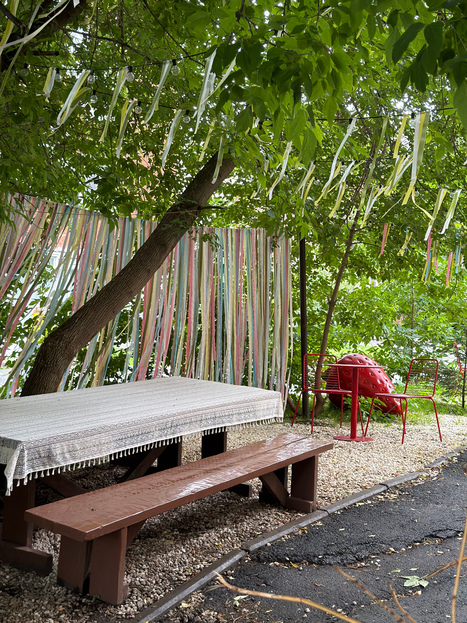 The image depicts an outdoor scene at what appears to be a park or garden area. The primary subjects are a picnic table with a textured cloth draped over it, and a decorative red strawberry-shaped object with accompanying red chairs, set against a backdrop of lush green trees. Hanging from the branches of the trees are numerous colorful ribbons of various shades, including pastel pink, yellow, green, and white, interspersed with small, clear spherical ornaments that could be lights or decorative beads. The ribbons create a festive and whimsical atmosphere.
The picnic table is made of dark brown wood and has visible bench seating. The cloth on the table is light-colored with intricate patterns and fringed edges. The red strawberry sculpture and chairs are a prominent visual element, adding a playful touch to the setting. The ground around the table and sculpture is covered in small, light-colored gravel. To the right of the gravel area, a dark asphalt path is visible, bordered by a dark gray strip.
The scene is brightly lit, suggesting it is daytime, possibly mid-morning or afternoon, under clear or partly cloudy skies. The abundance of green leaves indicates a season of growth, likely spring or summer. The overall mood conveyed by the image is cheerful and inviting, possibly suggesting an outdoor gathering, celebration, or a pleasant recreational space. There is no visible text in the image. The location context provided is Perm, Russia.