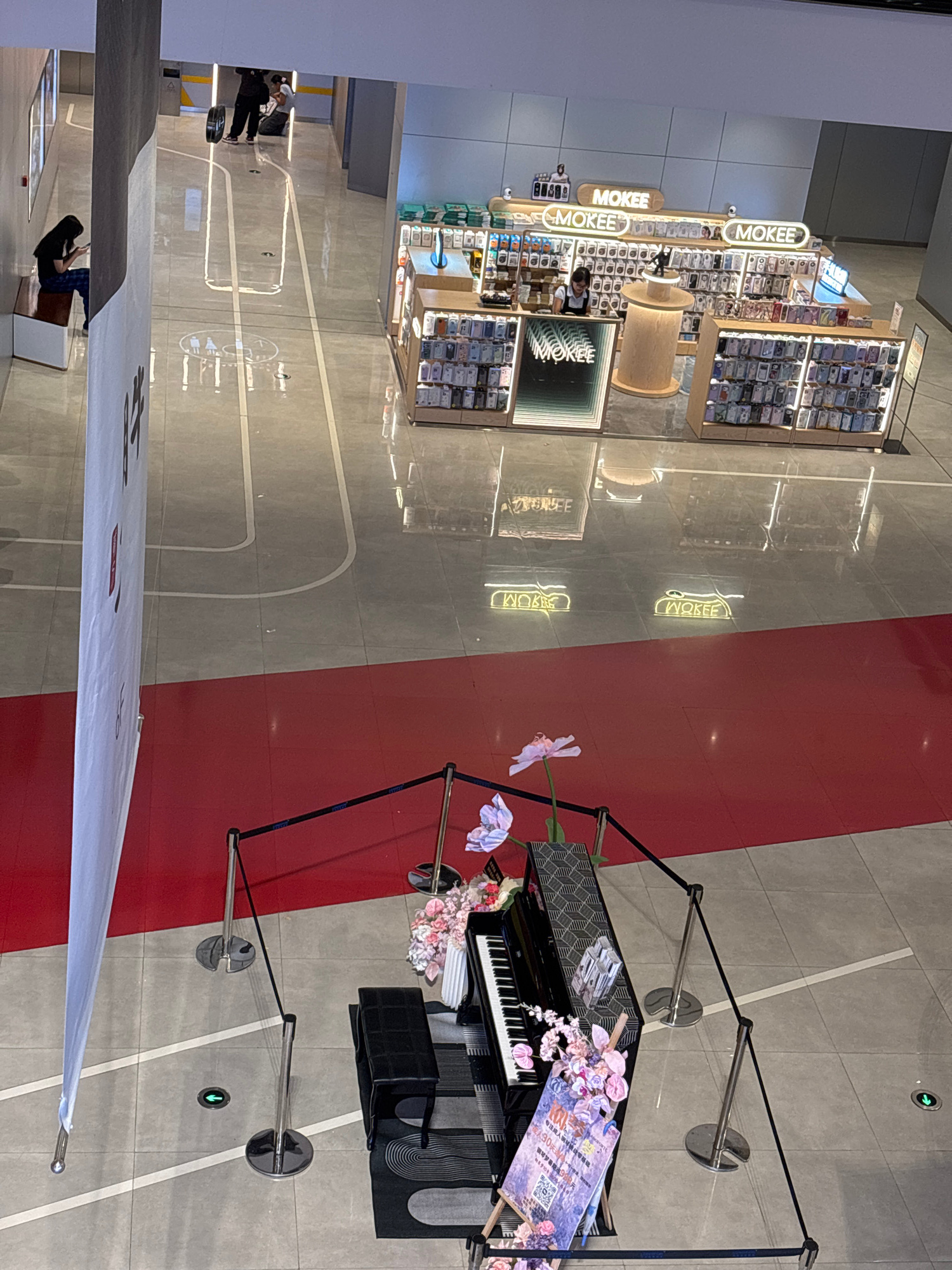 This image, taken from an elevated perspective, captures a section of a modern, indoor commercial space, possibly a shopping mall, in Shapingba District, China. The floor is a light gray tile with white line markings that suggest a pathway or sports court. A red carpeted area is visible in the foreground, separated from the tiled area by a low black rope barrier supported by chrome stanchions.
In the foreground, on the red carpet, is a grand piano with a black bench. The piano is decorated with floral arrangements in shades of pink and white. A sign with Chinese characters and a QR code is propped against the piano.
Further back, beyond the red carpet and across a tiled expanse, is a retail kiosk or store named "MOKEE". The store is brightly lit and displays a wide array of phone cases on shelves. Two individuals are visible behind the counter, and the store has illuminated signage.
In the background, on the tiled floor, there is a person sitting on a bench, looking at their phone. Another group of people are seen walking in the distance. The overall environment appears clean and well-lit, with a contemporary aesthetic. Reflections are visible on the polished floor, indicating a smooth, possibly glossy surface.