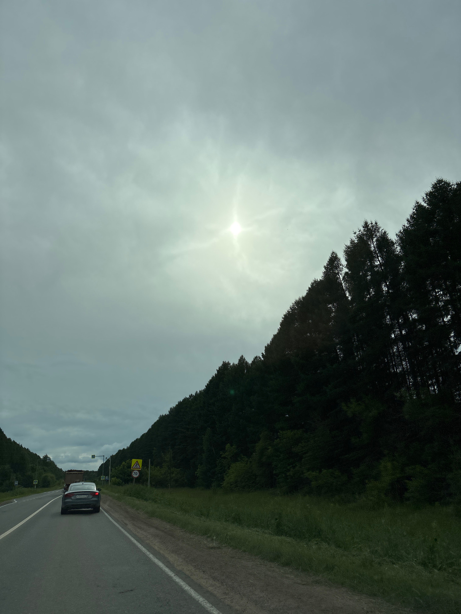 The image captures a view from a moving vehicle on a paved road through a wooded area, likely in Забегалово, Russia. The road curves to the left, with a white line marking the lane separation. A dark-colored sedan is driving ahead in the right lane. On the right side of the road, a steep incline is covered by dense, dark green evergreen trees. The sky above is overcast with a bright sun peeking through, creating a diffused light. Several yellow triangular road signs with black symbols are visible on the right side of the road, one with a number "70" indicating a speed limit. On the left side of the road, there are also smaller yellow road signs with black symbols. The terrain suggests a rural or semi-rural setting.