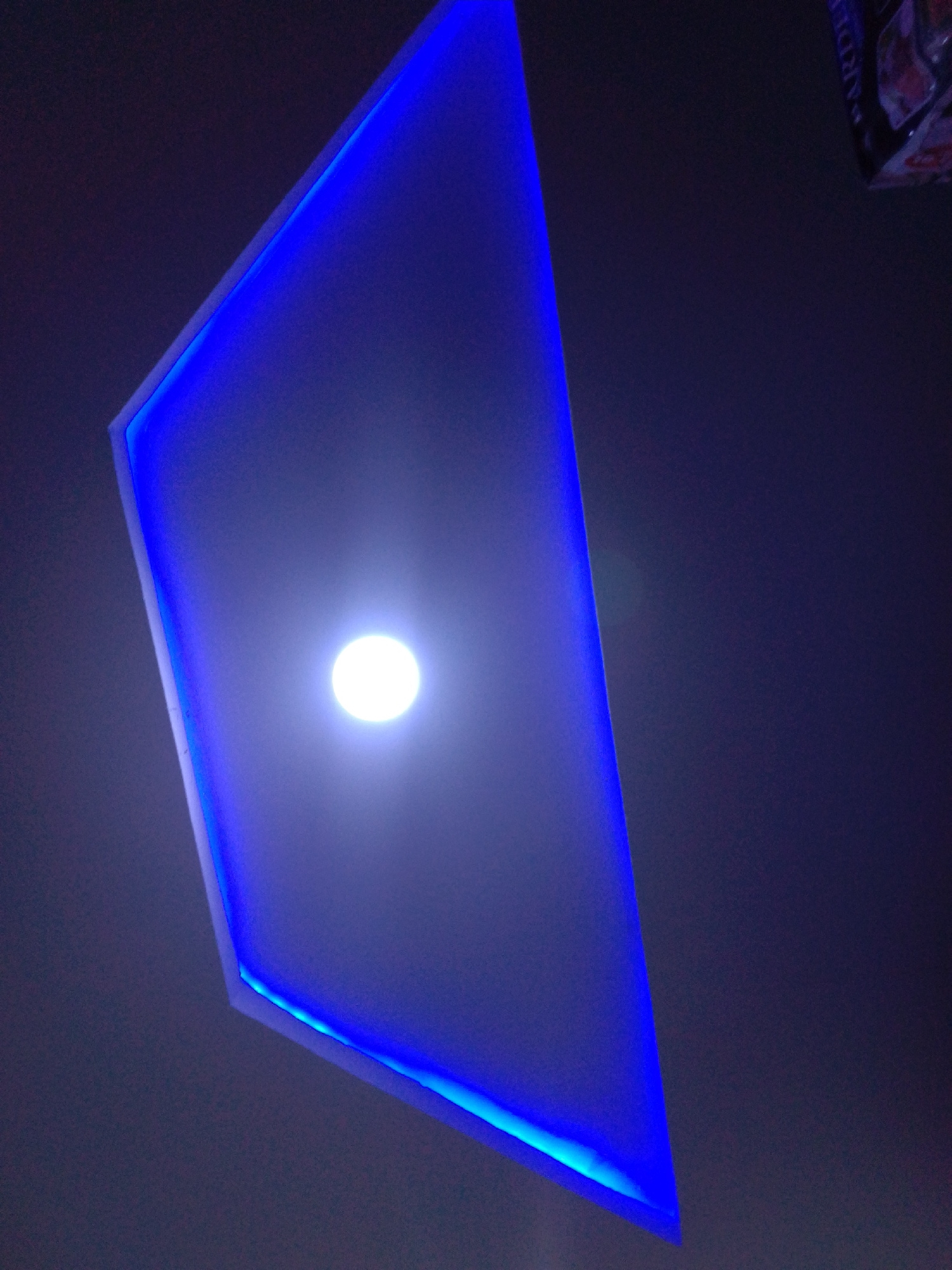 The image shows a ceiling with a modern, angular design featuring blue LED lighting along its edges. A bright, circular light source is visible at the center of the ceiling. The overall color palette is dominated by dark blues and grays, with the bright central light and the vibrant blue edge lighting creating a striking contrast. The image appears to be taken from below, looking upwards. There is no visible indication of the city of Howayi, Nigeria, or any specific location within it. No people, actions, or interactions are depicted in the image. The scene is static and focuses on the architectural lighting design.