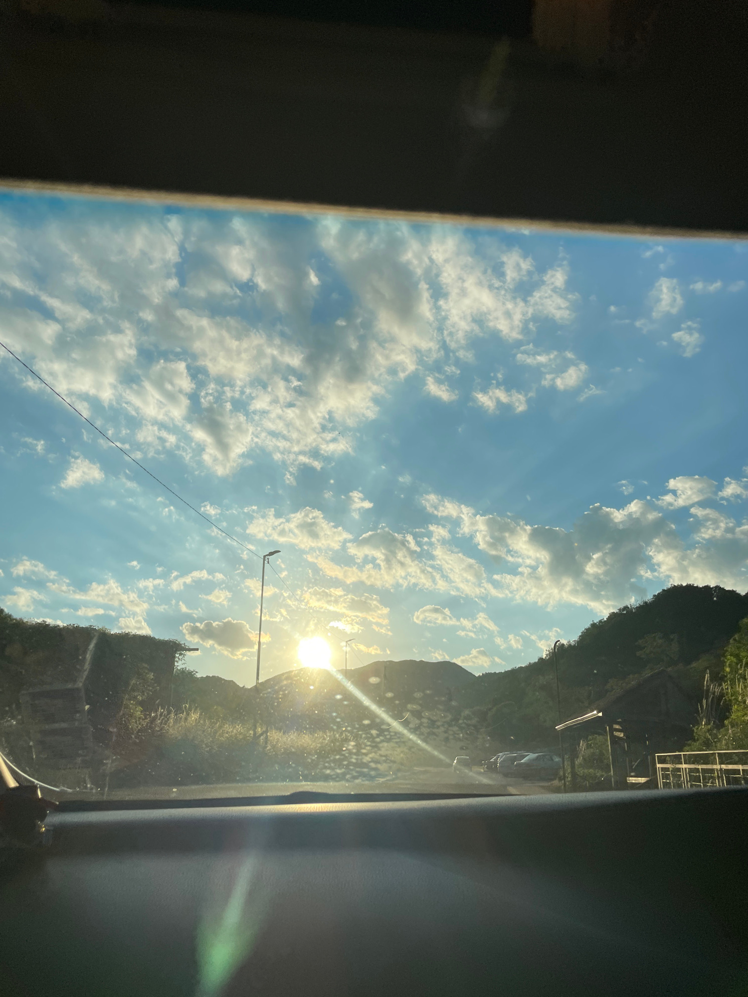 The image shows a view from inside a vehicle looking out through the windshield. The sun is low in the sky, casting long rays through scattered clouds, creating a dramatic sky with beams of light. The landscape appears to be mountainous and rural, with lush green vegetation and trees on the hillsides. In the foreground, a dark dashboard is visible, and the windshield has numerous water spots or dust particles, some of which are illuminated by the sun.
A street lamp is visible on the right side of the road, and there appears to be a parking area with several cars. On the left side, a building with a vine-covered wall is present. A rustic wooden structure, possibly a pavilion or shelter, is situated on the right side of the road, along with a metal railing. The overall impression is a scenic, possibly a sunset or sunrise view, in a natural environment.
Based on the available information, this image could be from the vicinity of Zlatograd, Bulgaria, a town known for its preserved Revival-era architecture and proximity to natural landscapes.