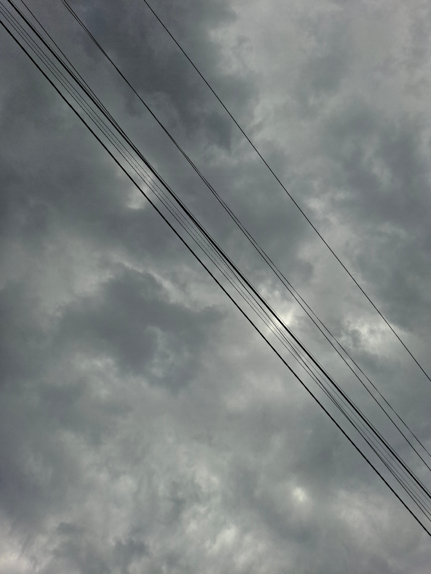 The image captures a close-up, angled view of multiple parallel black electrical wires against a heavily overcast, gray sky. The wires are positioned diagonally from the bottom left to the upper right of the frame, with varying degrees of spacing between them. The sky is filled with dense, textured clouds, suggesting a gloomy or stormy weather condition. There are no people, structures, or other distinguishing features visible in the frame, making the wires and the sky the sole subjects of the photograph. The scene appears to be an outdoor environment, likely in an urban or semi-urban setting given the presence of electrical infrastructure. The specific location in Төмөнкү Ала-Арча, Kyrgyzstan is not visually discernible from the image itself, but the context implies the presence of such utility lines in that area.