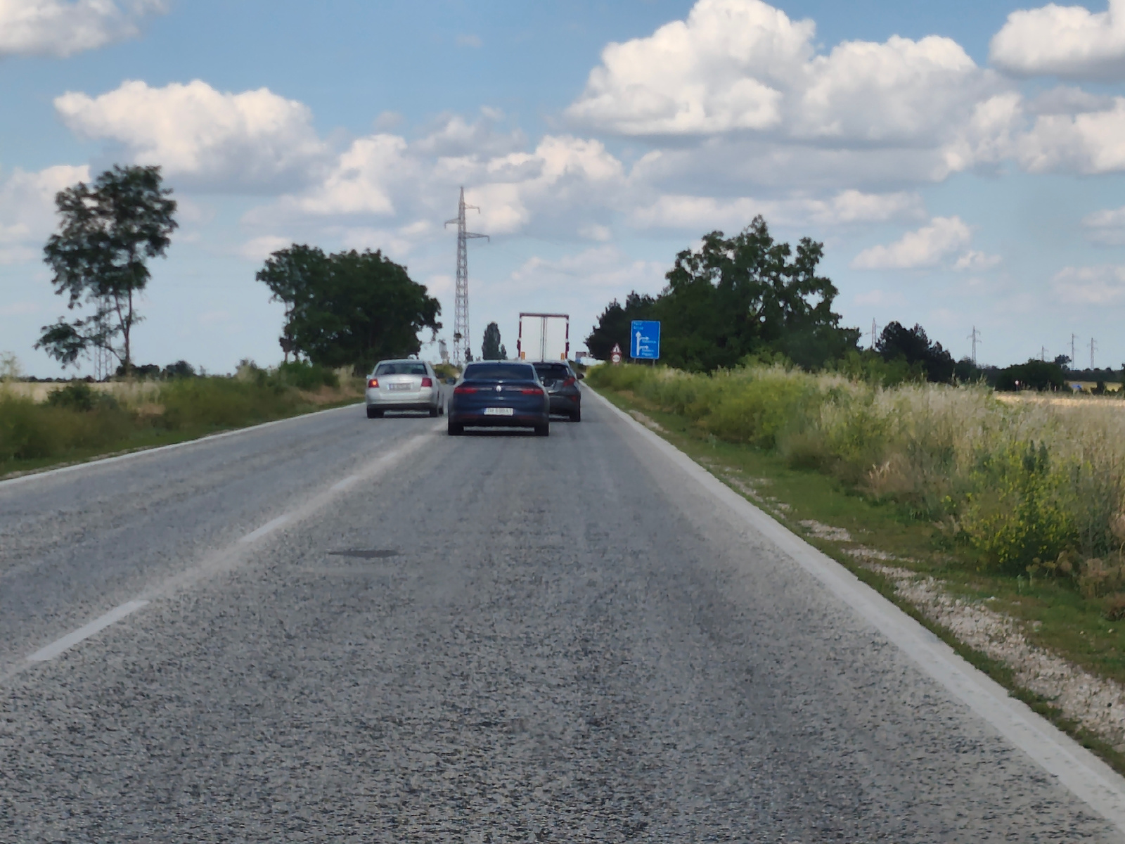 The image shows a view from a moving vehicle on a paved road with a white dashed line on the left side. The road is wet in some areas, suggesting recent rain. The sky is bright blue with fluffy white clouds, indicating a daytime scene, likely late morning or early afternoon.
Several cars are visible on the road ahead. The closest car in the right lane is dark blue. Further ahead, a white car is in the left lane, and a dark car is in the right lane. Beyond them, a white truck is visible.
The landscape on either side of the road consists of grassy fields and scattered trees. There are also electricity pylons in the distance. On the right side of the road, a blue sign with white symbols is visible, indicating directions.
The scene appears to be a rural or semi-rural area. The presence of a highway and multiple vehicles suggests a journey or commute. There are no people clearly visible in the image, only the cars and the environment. The overall mood is neutral, depicting a typical drive on a cloudy day. The image is not of high resolution, so fine details are not discernible.