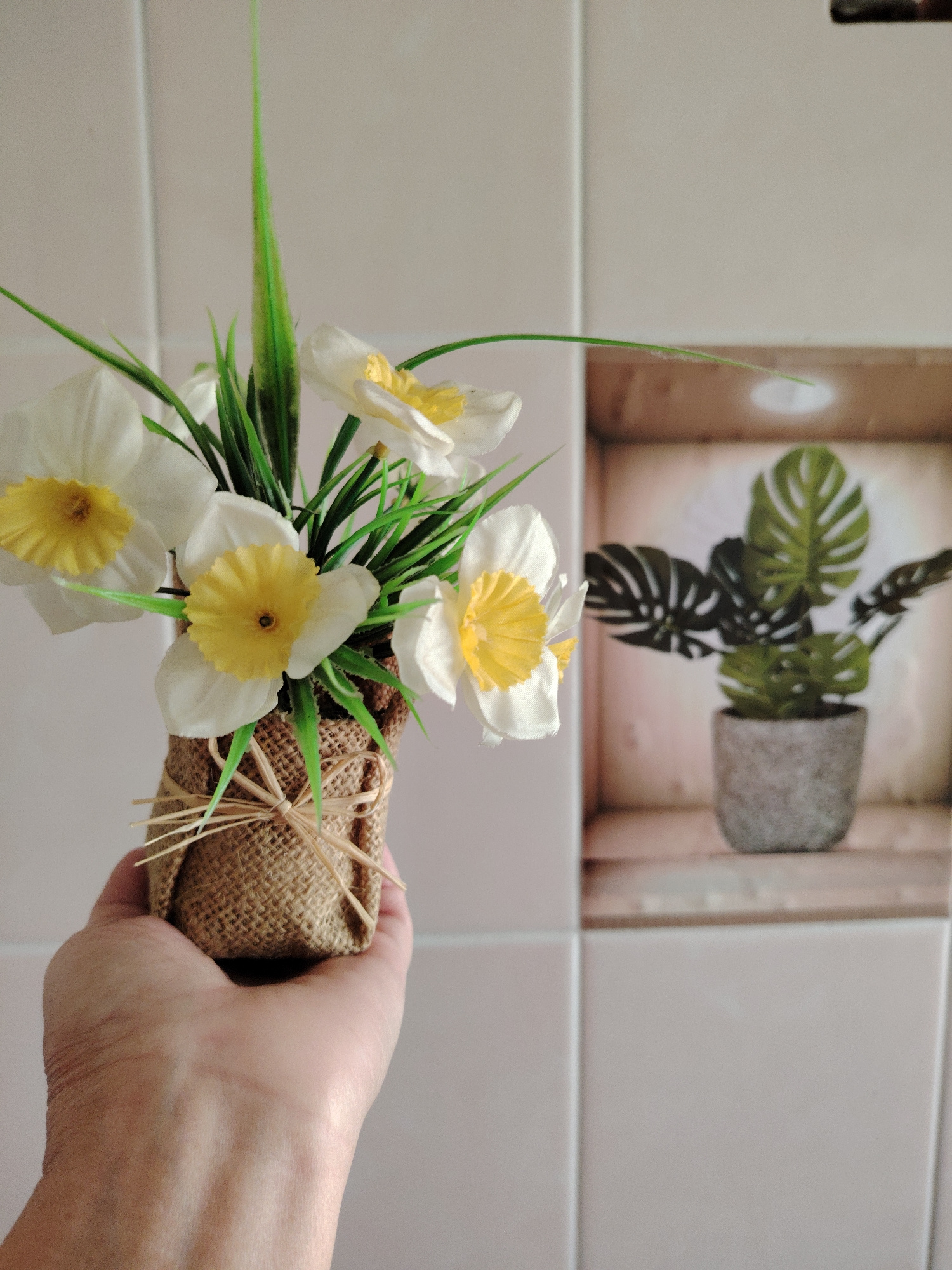 A hand holds a small burlap-wrapped pot containing artificial white daffodils with yellow centers and green leaves. In the background, a wall with square tiles features a decorative insert depicting a potted Monstera plant in a concrete pot. The scene is illuminated with soft, diffused light. The location is not specified in the image and cannot be determined to be Soivolovskaya, Russia.