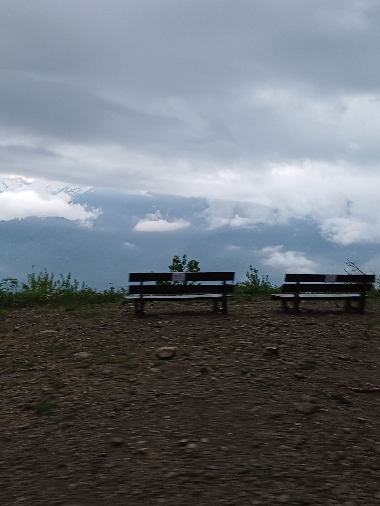 The image depicts a scenic mountain overlook in Estosadok, Russia, featuring two dark wooden park benches positioned on a gravelly path. The benches are oriented towards a vast landscape of layered, mist-shrouded mountains under a dramatic, cloudy sky. The foreground is dominated by the rough texture of the path, with scattered stones and sparse green vegetation. In the background, the mountains rise in successive planes, with some peaks appearing snow-capped, while others are obscured by soft, grey clouds. The overall atmosphere is serene and somewhat dramatic due to the overcast sky and the imposing mountain scenery. There are no people visible in the image.