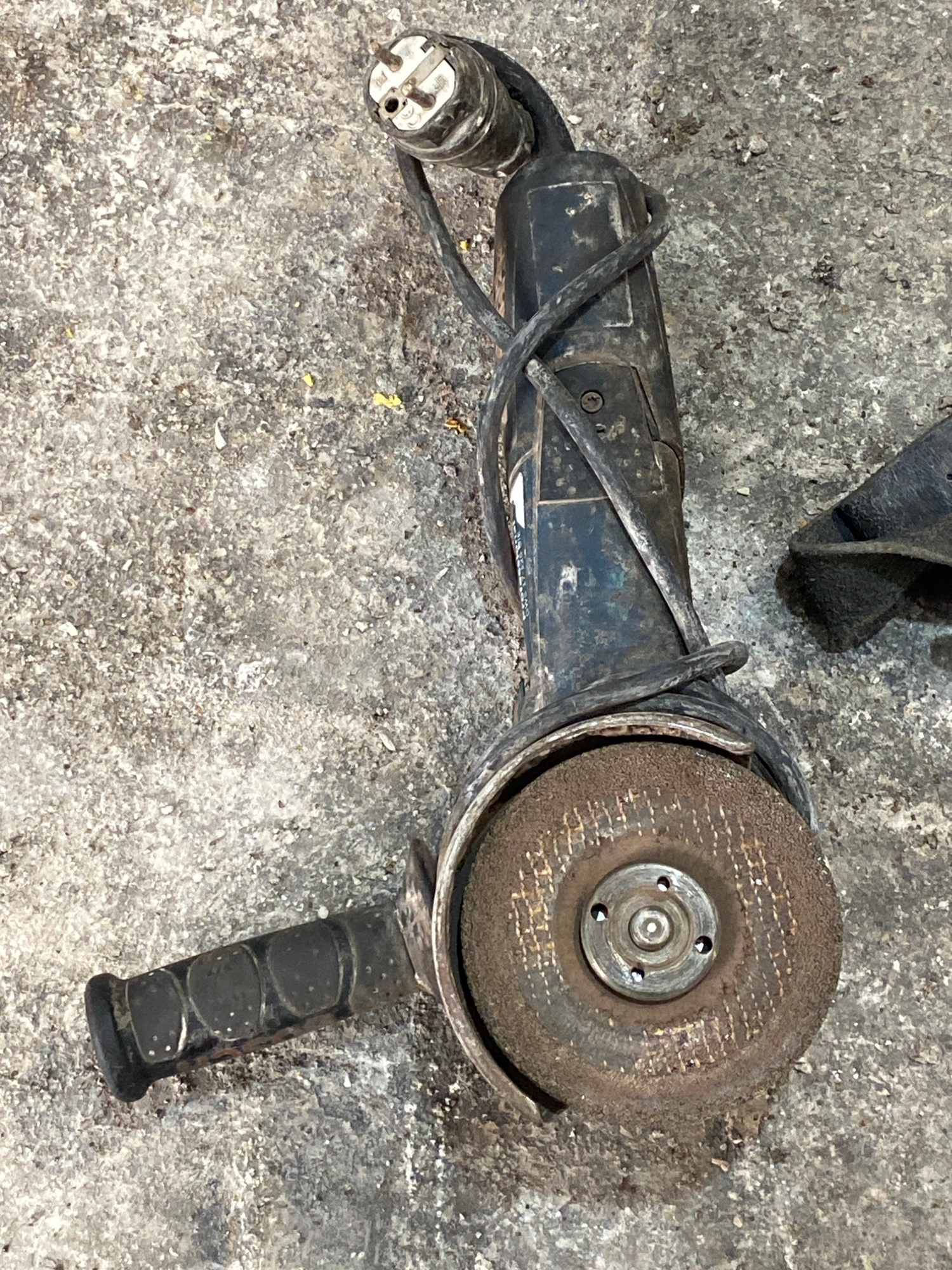 The image shows an angle grinder lying on a rough concrete or asphalt surface. The angle grinder is black and appears to be used, with dust and grime on its body and the grinding disc. The power cord is wrapped around the tool. A black auxiliary handle is attached to the side. To the right of the angle grinder, a piece of black material, possibly a glove or another tool accessory, is visible. The overall scene suggests a workshop or outdoor work area. The location in the city of Police, Poland is not discernible from the image.