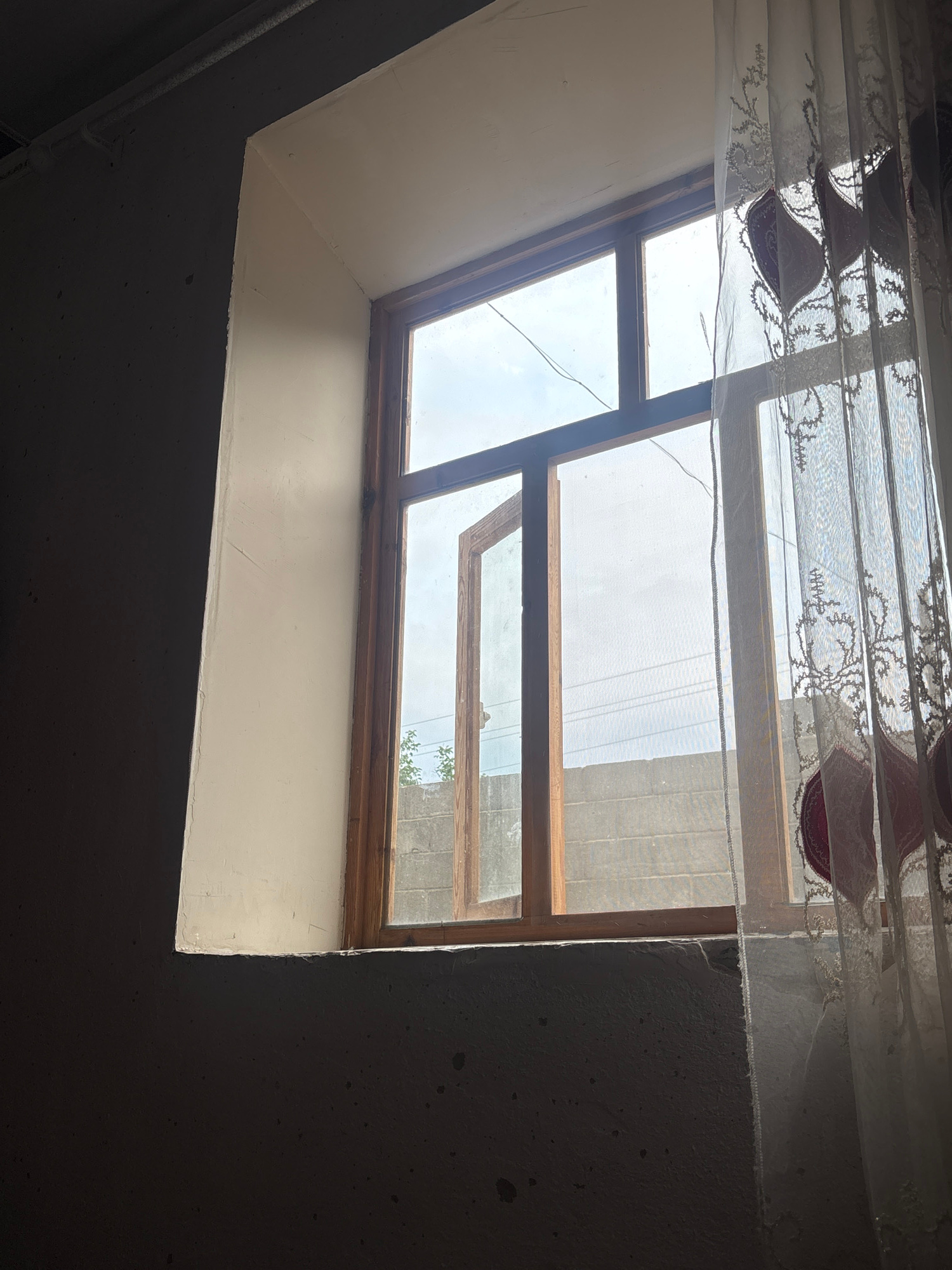 A close-up, low-angle shot captures a simple window with wooden frames, set within a rough, dark-colored wall. The window is partially open, revealing a view of a dull, overcast sky and a gray cinder block wall outside. A sheer, white lace curtain with a floral pattern hangs on the right side of the window, partially obscuring the view and casting shadows. The interior wall surrounding the window is painted a light cream color. The overall lighting is dim, with light streaming in from the window, creating a stark contrast between the illuminated window area and the shadowed foreground. The scene appears to be indoors, possibly in a residential or utilitarian space.