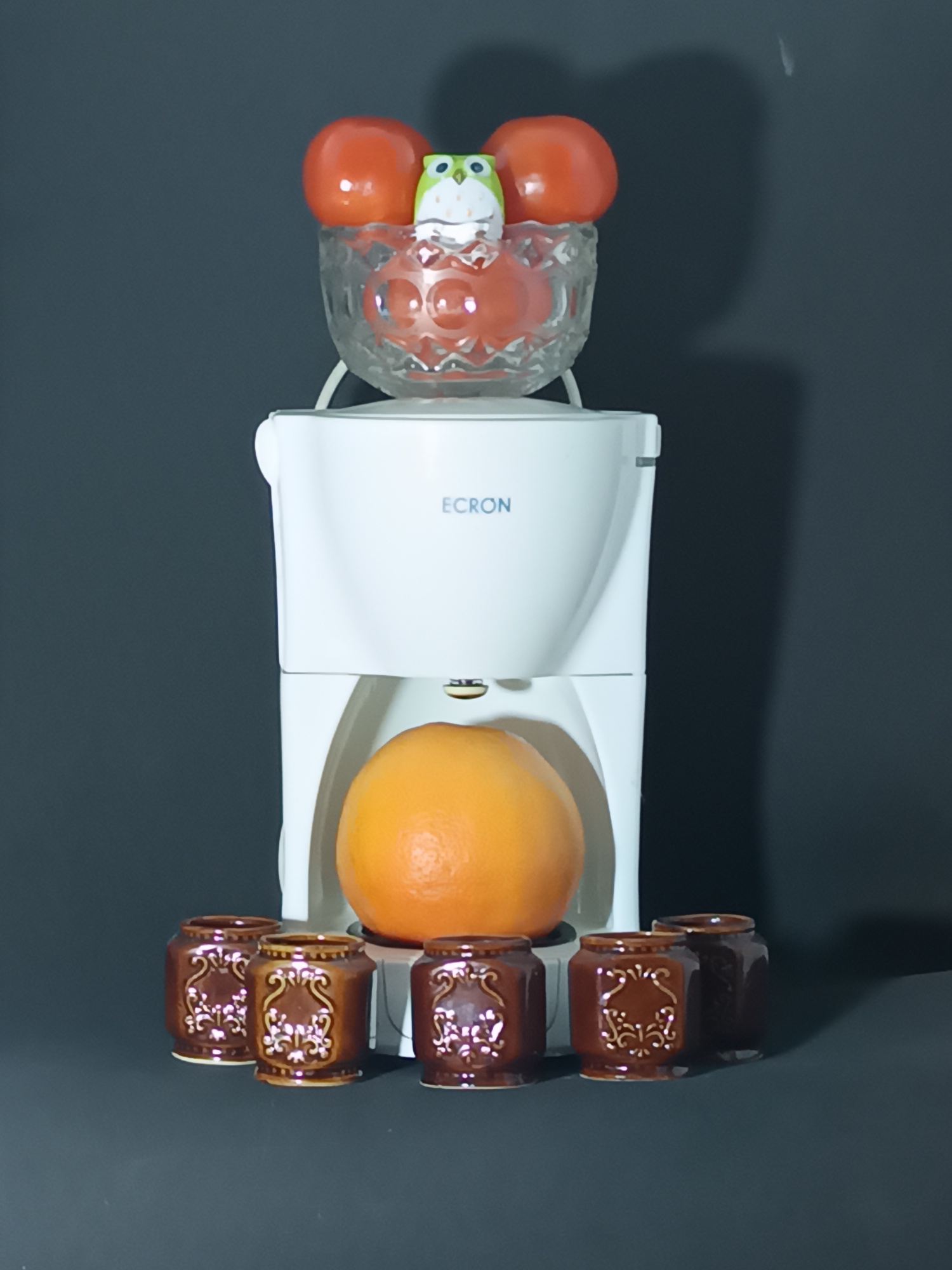 A still life arrangement features a white coffee maker as the central object, with a glass bowl filled with red items and a small, colorful owl figurine perched on top. Below the coffee maker, a large orange rests in a cradle. Arranged in front of the coffee maker are several small, brown ceramic containers with decorative patterns. The background is a plain, dark gray, suggesting a studio setting or a room with minimal ambient light. The lighting creates subtle shadows, highlighting the textures and shapes of the objects. The scene is devoid of people, and no specific activity is taking place; it is a static composition. The text "ECRON" is visible on the coffee maker. The setting does not offer strong cues for time of day or weather, and no emotional tones are conveyed by the arrangement. There are no explicit location cues within the image itself.