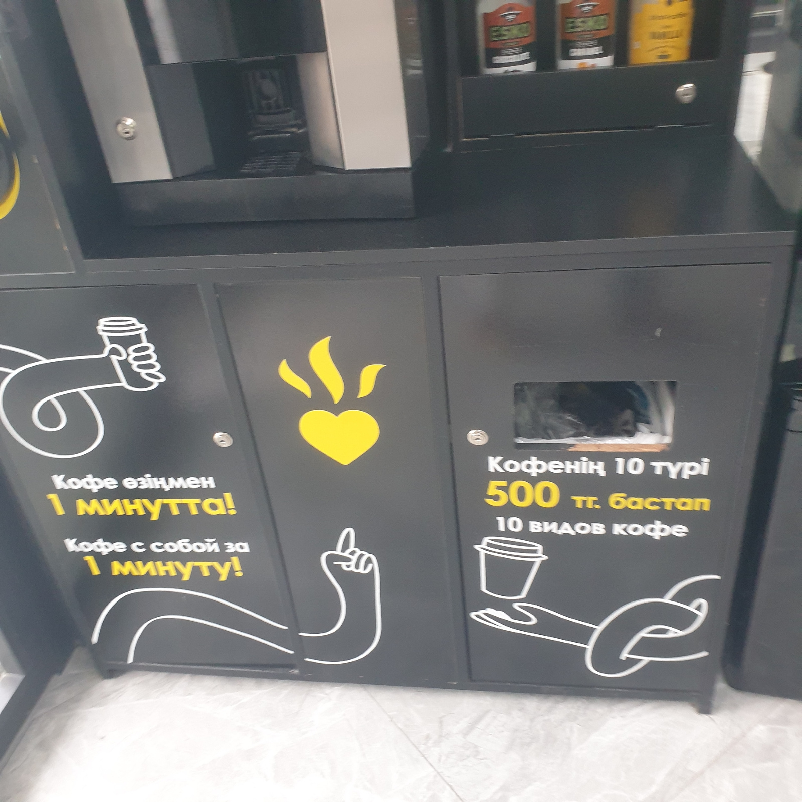 The image depicts a coffee vending machine with Cyrillic text. The machine has a black exterior with a gray upper section. On the left side of the machine, there is a white graphic of a hand holding a coffee cup, with the text "Кофе өзiңмен 1 минутта!" (Coffee with you in 1 minute!) and "Кофе с собой за 1 минуту!" (Coffee to go in 1 minute!) below it. On the right side, there is a yellow graphic of a heart with flames, and text that appears to be a price and description: "Кофенiң 10 түрi 500 тг. бастan 10 видов кофе" (10 types of coffee from 500 tenge, 10 types of coffee). There is a small window on the right side showing contents within the machine. The machine is positioned on a light-colored tiled floor. The overall impression is of an advertisement or information display for a coffee service. The specific location in Shymkent, Kazakhstan, cannot be determined from the image alone, but the presence of Cyrillic text strongly suggests it is within Kazakhstan or a neighboring country with a significant Russian-speaking population.
