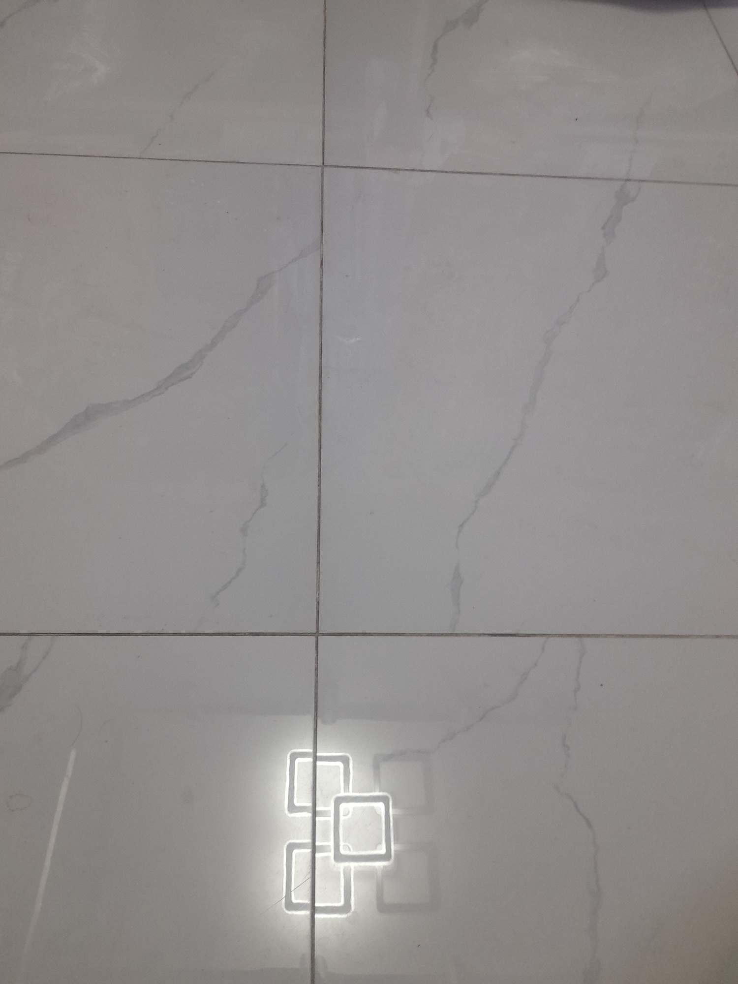 The image displays a floor covered in large, rectangular white tiles with subtle gray veining, resembling marble. Four tiles meet to form a central point in the frame. In the bottom right quadrant of the image, a bright, stylized reflection of interconnected square outlines is visible on the glossy surface of the tiles. This reflection suggests a light source, possibly a modern decorative light fixture, positioned above and reflecting onto the floor. The overall scene is clean and minimalist, focusing on the texture and reflectivity of the tiled surface. The location is the city of Unknown, Unknown.