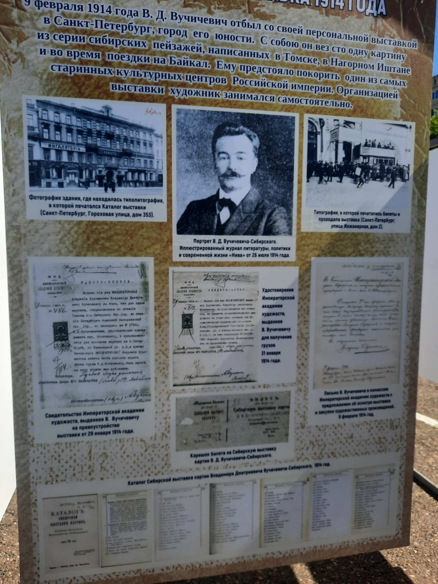 The image shows a vertical informational display board, likely situated outdoors in a park or public area in Zelenogorsky, Russia. The board contains text and photographs detailing the life and work of V. D. Vuchichevich, likely an artist or cultural figure. The top of the board features a title in large, bold font, partially obscured but containing the year "1914". Below this, descriptive text in Russian elaborates on Vuchichevich's personal exhibition, his travels, and his involvement in cultural events.
The board is divided into sections, each with accompanying photographs and text. One section displays a black and white photograph of a multi-story building with architectural details and signage that reads "ИНГАЛЯТОРІН". The caption below this photograph, in Russian, identifies it as the building where the exhibition catalog was printed. Another section features a portrait of a man with a mustache, identified as V. D. Vuchichevich. Below the portrait is text mentioning the journal "Niva" and a date in July 1914.
Other sections showcase what appear to be official documents, such as a "Certificate of the Imperial Academy of Arts" and a letter from Vuchichevich to a commission. There are also reproductions of exhibition tickets and catalog pages, with lists of artworks. The overall impression is of a historical exhibit dedicated to an artist and his significant exhibition in 1914. The background of the display board is a textured, light brown color, and the edges of the board are visible, suggesting it is a freestanding unit. The surrounding environment is blurred, but hints of greenery and possibly sky suggest an outdoor setting.
