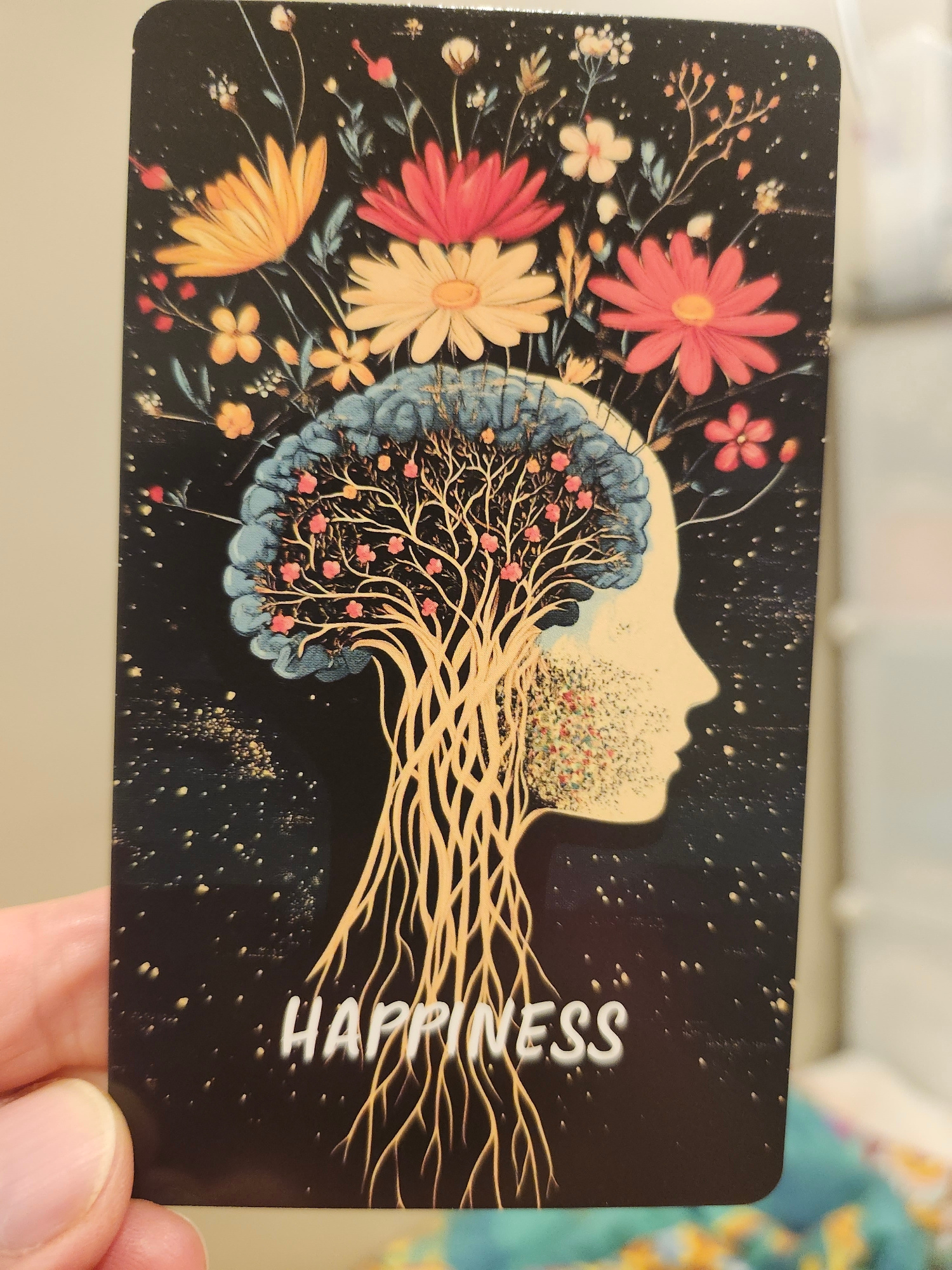 A hand is holding a rectangular card against a blurred background. The card features an illustration of a silhouette of a human head in profile, facing right. The head is depicted as a tree with roots extending downwards and branches growing upwards into a cluster of flowers. The tree's trunk and branches are light brown, and small pink flowers are interspersed within the branches. The top of the head is adorned with various colorful flowers in shades of yellow, pink, and orange, set against a black, starry background. The word "HAPPINESS" is written in white, stylized lettering at the bottom of the card. The background of the card is black with small white and gold speckles, resembling stars. The hand holding the card is light-skinned and appears to be part of an adult. The blurred background suggests an indoor environment, possibly a room with muted lighting and other objects out of focus. There is no discernible location in the city of Gatineau, Canada, visible in the image.
