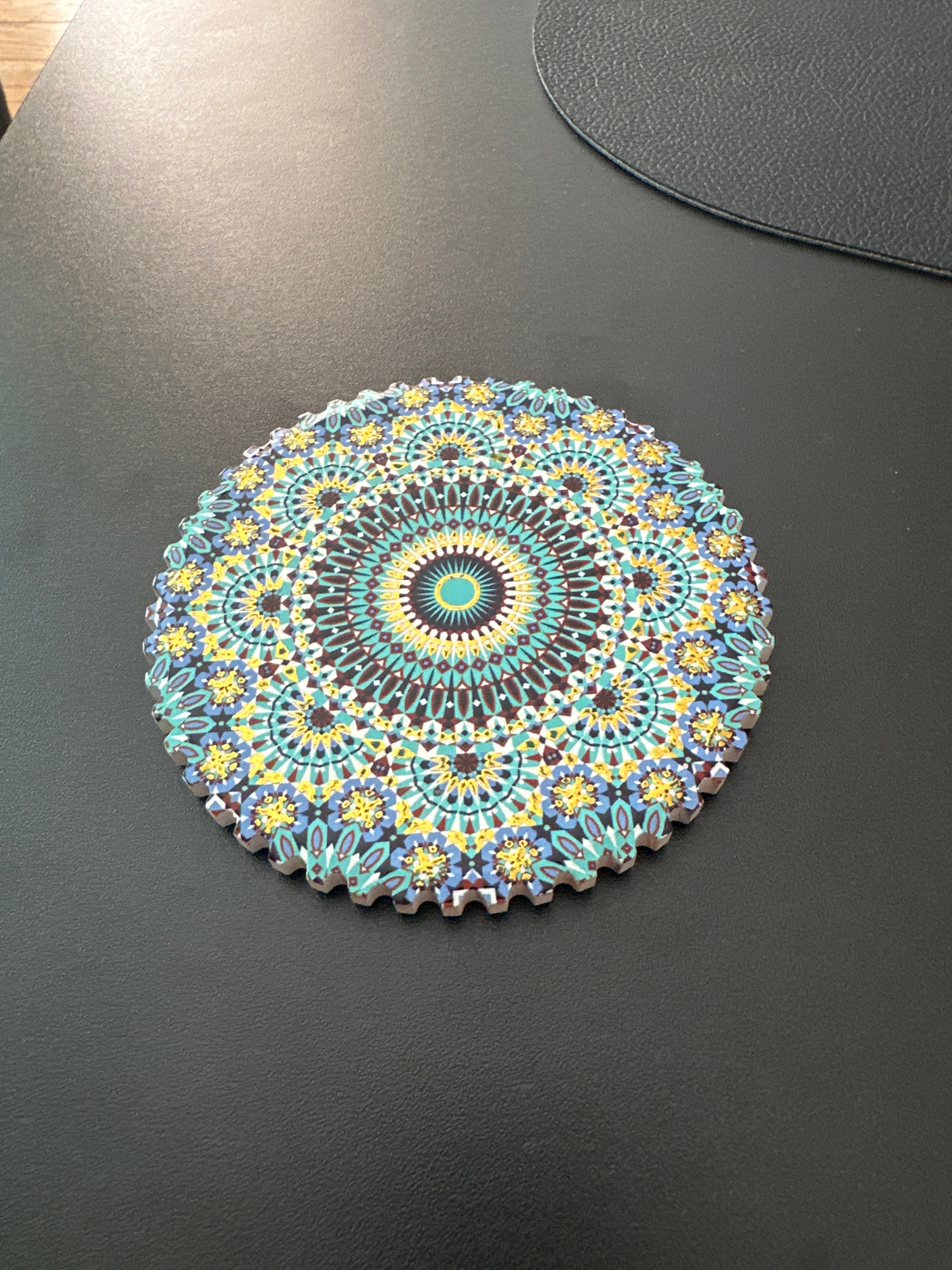 A circular coaster with a colorful mandala pattern is placed on a dark, textured surface, possibly a desk or table. The coaster features intricate geometric designs in shades of blue, yellow, teal, and brown, with a bright turquoise center. The edge of the coaster is serrated. A dark gray, rounded object, possibly a mousepad, is visible in the upper right corner of the frame. The lighting suggests an indoor environment. There are no people or animals present in the image. The location is not specified as Warsaw, Poland.