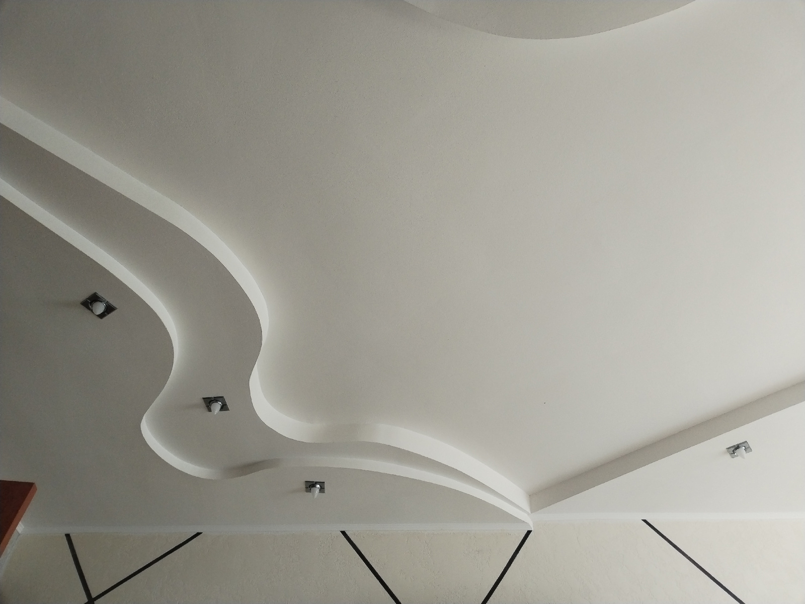 The image shows a white, modern false ceiling with decorative curves. Recessed lighting fixtures are visible on the ceiling. The lower portion of the image shows a wall with a textured finish and black linear decorative elements. The scene is an interior view, likely a room. The location of this scene within Pastavy, Belarus, is not discernible from the visual information provided.