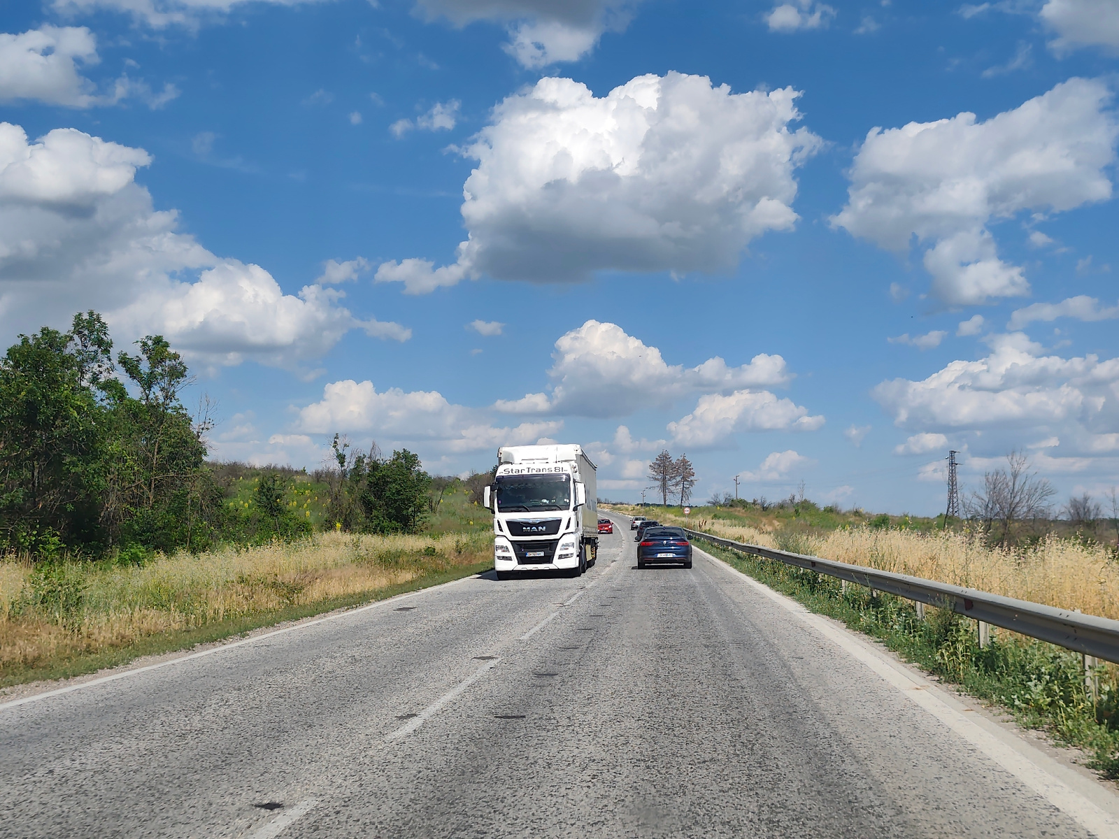 A forward-facing, medium-shot, eye-level, wide-angle perspective captures a sunny day on a paved road in Pleven, Bulgaria. The road, textured with a gray, pebbled surface, is marked with white lines and has occasional dark patches. On the right side of the road, a metallic guardrail runs along a grassy verge. The landscape to the left and right is characterized by dry, yellow grass and scattered green bushes and trees, with a few bare trees visible in the distance. The sky is a bright blue, dotted with fluffy white clouds.
In the scene, a white semi-truck with the branding "Star Trans B." and "MAN" on its front, is driving on the road. The truck has a partially obscured license plate. Ahead of the truck, a dark blue sedan is also traveling in the same direction. Several other vehicles are faintly visible further down the road. A distant power pylon stands against the horizon. The overall scene suggests a journey on a clear, warm day.