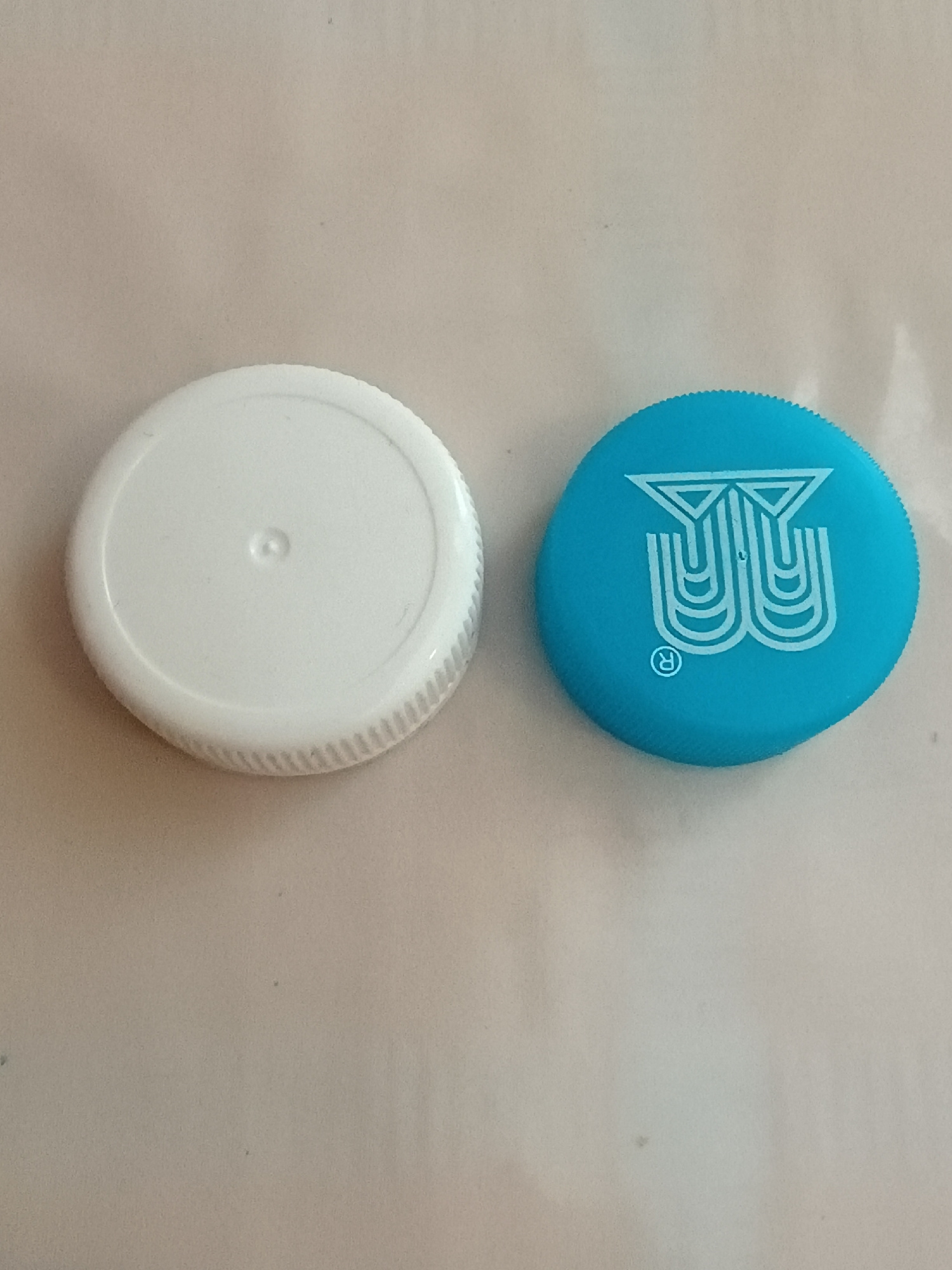 Two bottle caps are displayed on a light-colored, textured surface. The upper cap is a vibrant blue and features a stylized white "B" logo with a registered trademark symbol to its right. The lower cap is white and has a ribbed edge and a slightly raised circular indentation in its center. The lighting suggests an indoor setting, and there are no discernible environmental cues to indicate the time of day or weather. The surface appears to be a fabric or paper material, possibly a tablecloth or a desk mat. The objects are the primary focus, and there is no indication of any people or specific activities taking place. The image provides no information about the location context of Zaporizhzhia, Ukraine.