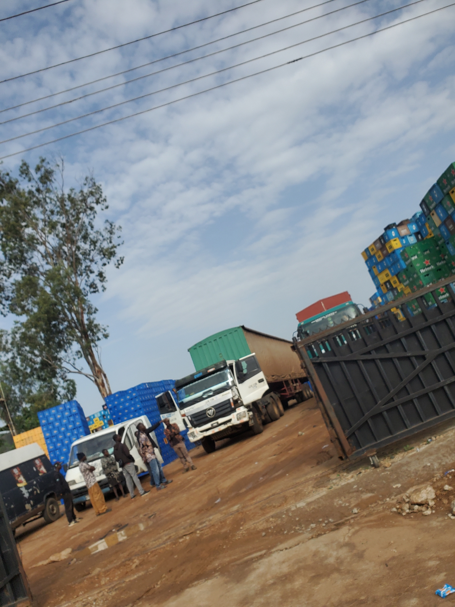 The image depicts an outdoor scene in Bukuru, Nigeria, possibly a distribution or loading yard. A fleet of vehicles is present, including a white delivery truck, a black van, and two larger trucks, one with a red trailer and another with a green cargo container. The vehicles are parked on a dirt ground with some puddles. Stacks of blue crates, possibly containing beverages, are visible behind some of the vehicles. In the foreground, a black metal gate stands open. Several individuals are interacting near the white delivery truck; one person is pointing, while others appear to be conversing. The sky is partly cloudy, and power lines are visible overhead. The overall impression is one of commercial activity, with goods being managed and transported.