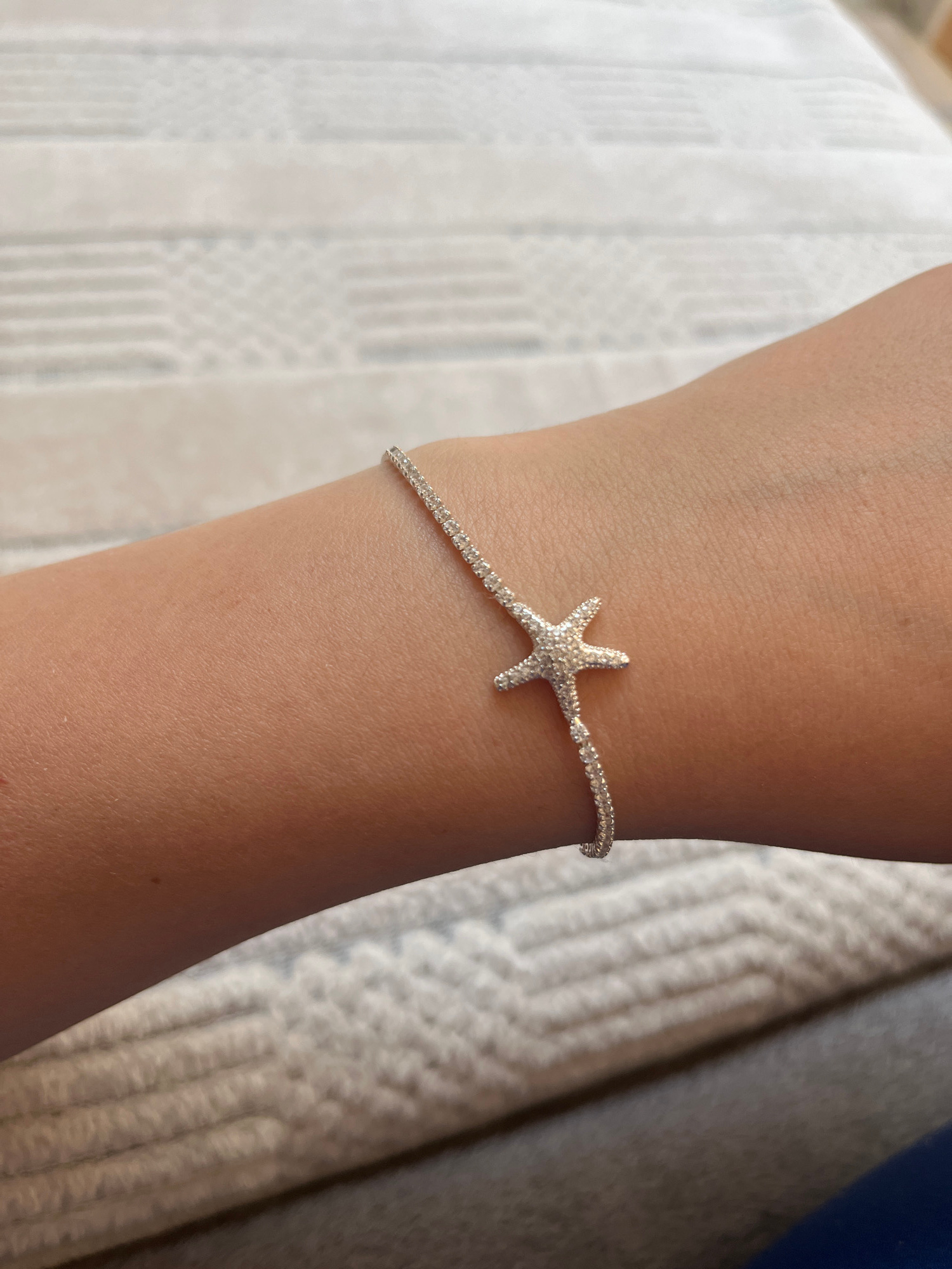 A close-up, high-angle shot shows a delicate silver bracelet with a starfish charm encrusted with small, clear stones on a person's forearm and wrist. The bracelet is positioned on the underside of the arm. The background is a textured, off-white surface with horizontal lines, possibly a blanket or upholstery. The lighting is soft and diffused. The image does not contain any elements or context that would place it within the city of Garmisch-Partenkirchen, Germany.