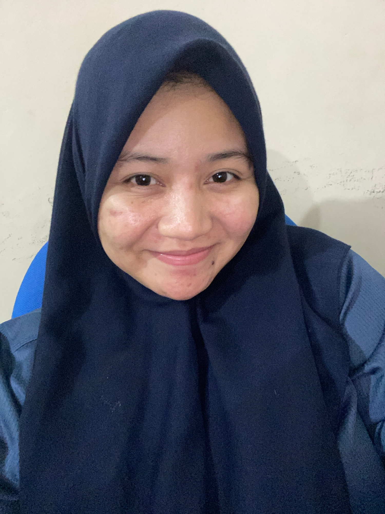 A close-up, medium shot shows a woman wearing a dark blue hijab and a blue collared shirt. She is looking directly at the camera with a slight smile. Her skin has some blemishes and redness on her cheeks and forehead. The background is a textured, light-colored wall. A portion of a blue chair is visible behind her left shoulder. The lighting suggests an indoor setting. The image appears to be a selfie taken in Banjarbaru, Indonesia.