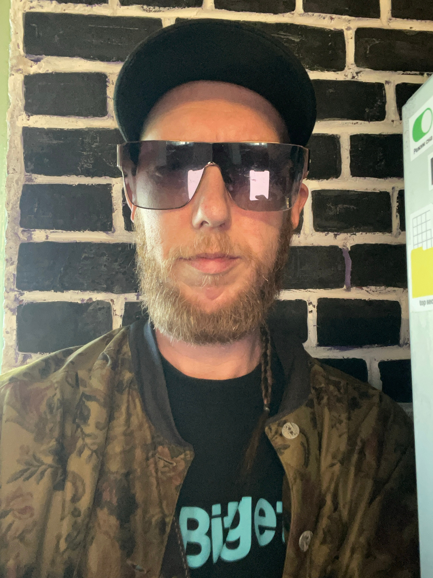 A man wearing a black flat cap, oversized sunglasses, a patterned jacket, and a black t-shirt with the word "Bigel" in blue letters, is standing in front of a brick wall. The wall is painted black with white mortar. To the right of the man, there is a white surface with some text and graphics. The lighting is natural and comes from the front. The man has a beard and a pigtail. He is looking directly at the camera. The overall scene is casual and appears to be indoors. There is no information within the image to determine the specific location within the city of Saint Petersburg, Russia.