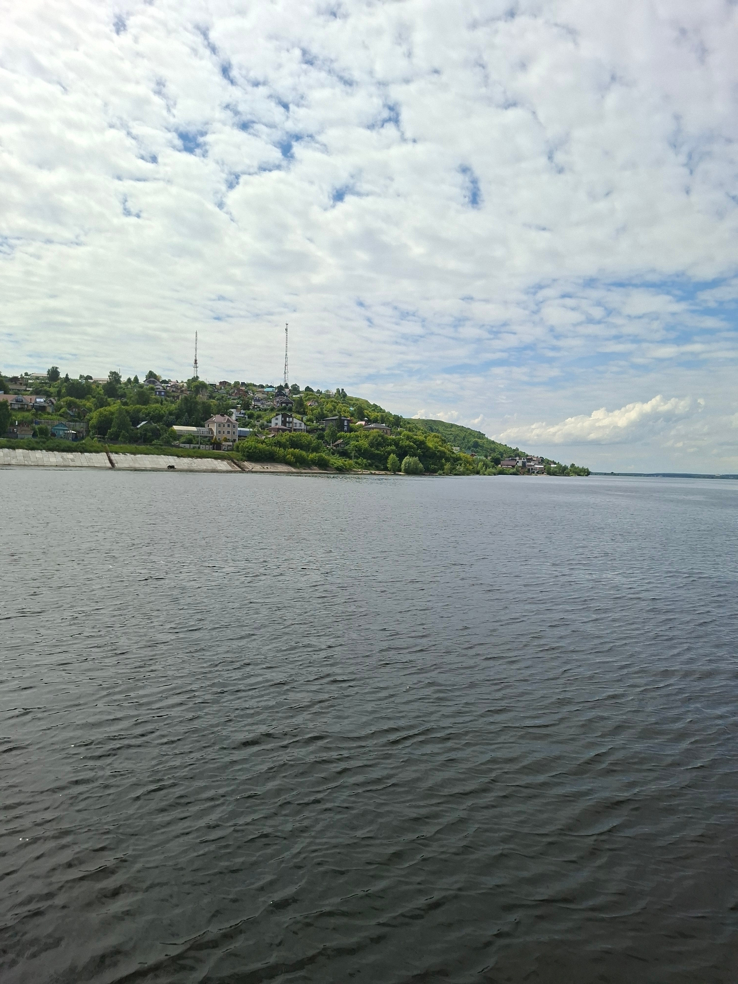 This is a landscape image captured from a side-on perspective, divided into two distinct halves: the sky and the water. The left half of the image is dominated by a cloudy blue sky. Fluffy white clouds are scattered across the expanse, with patches of blue sky visible between them. In the distance, along the horizon, a thin line of land is visible.
The right half of the image is a body of water, likely a river or lake, with a textured surface. The water is dark and appears to have ripples or small waves, suggesting a gentle breeze.
In the middle ground, along the shoreline, is a settlement situated on a gentle slope. Buildings with varying roof colors, interspersed with lush green trees, are visible, suggesting a populated area nestled amongst nature. Two tall communication towers are also present, breaking the natural skyline. The shoreline itself is lined with a concrete embankment.
The image appears to have been taken during the daytime, with the lighting suggesting it could be late morning or early afternoon. The presence of clouds indicates a partly cloudy day. The scene is tranquil and offers a view of a semi-rural or suburban setting by a large body of water. No text is visible in the image. The orientation of the image suggests it might have been taken with the camera held vertically.